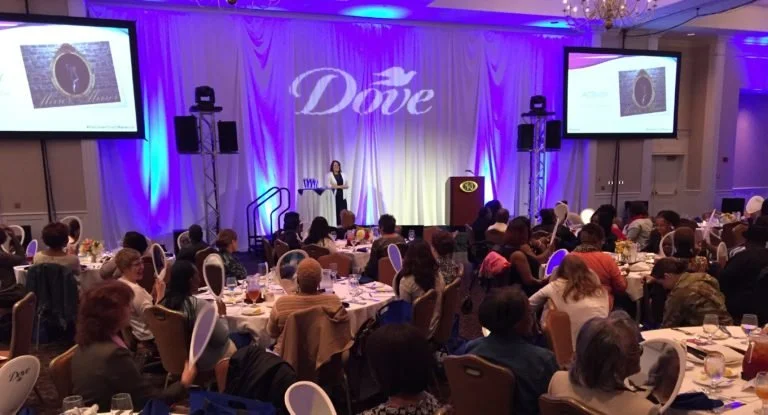 Veteran Caregivers seated at round tables in a banquet hall listening to speaker and military spouse caregiver Judy Davis on stage at a Dove Women's Conference