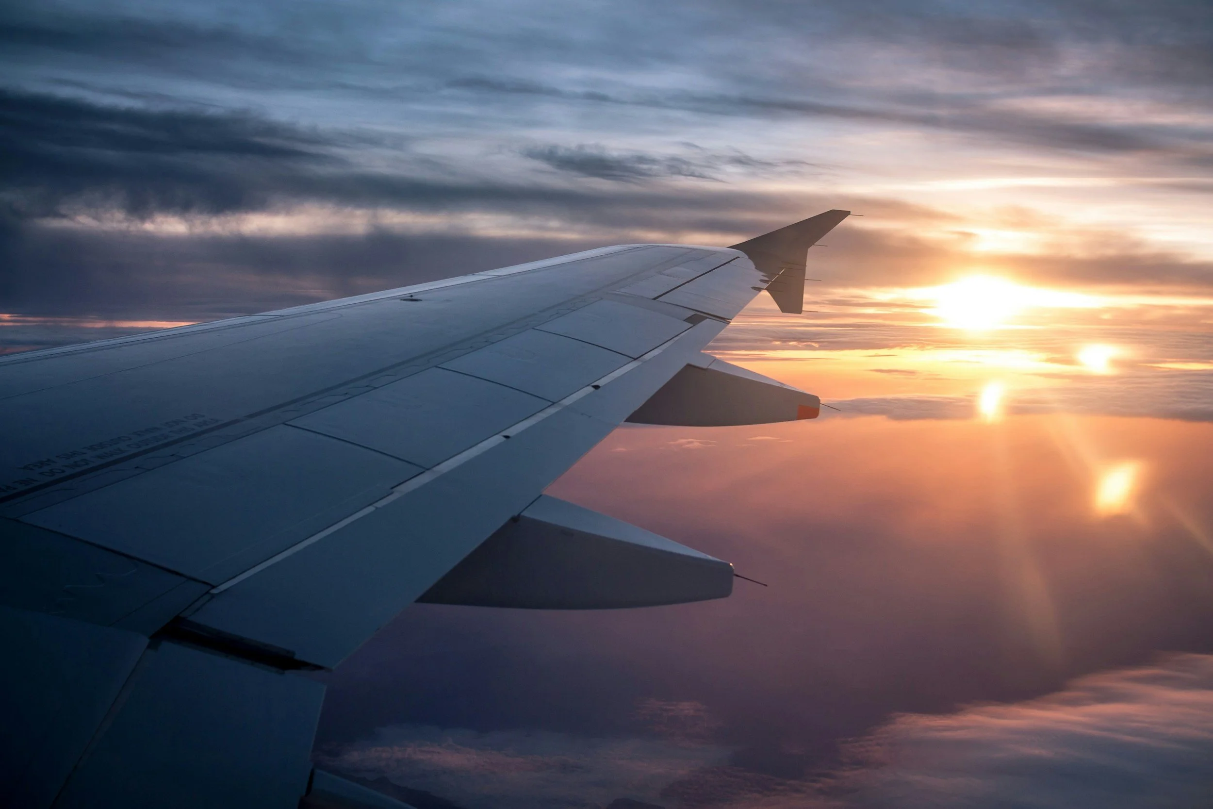 View from an airplane window showing airplane wing with the sun setting and clouds below.