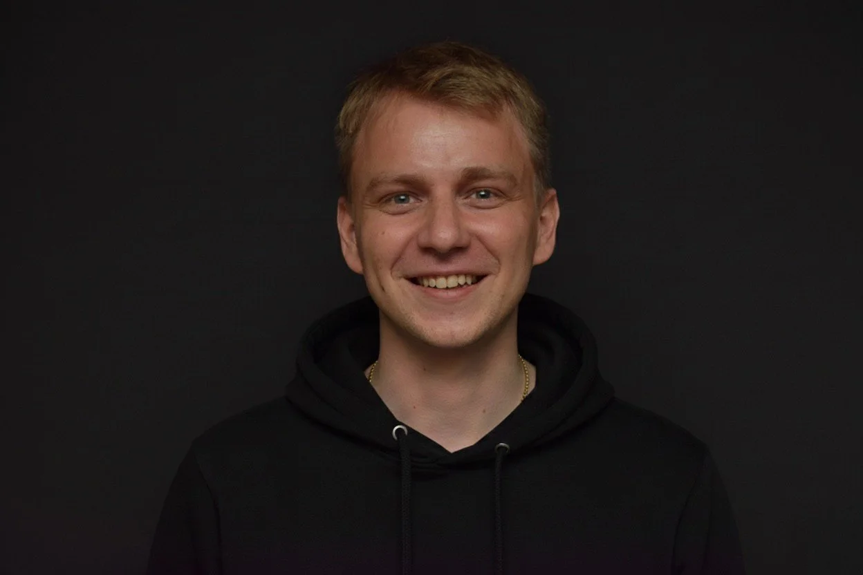 Young man with short blonde hair smiling, black hoodie, against a dark background.