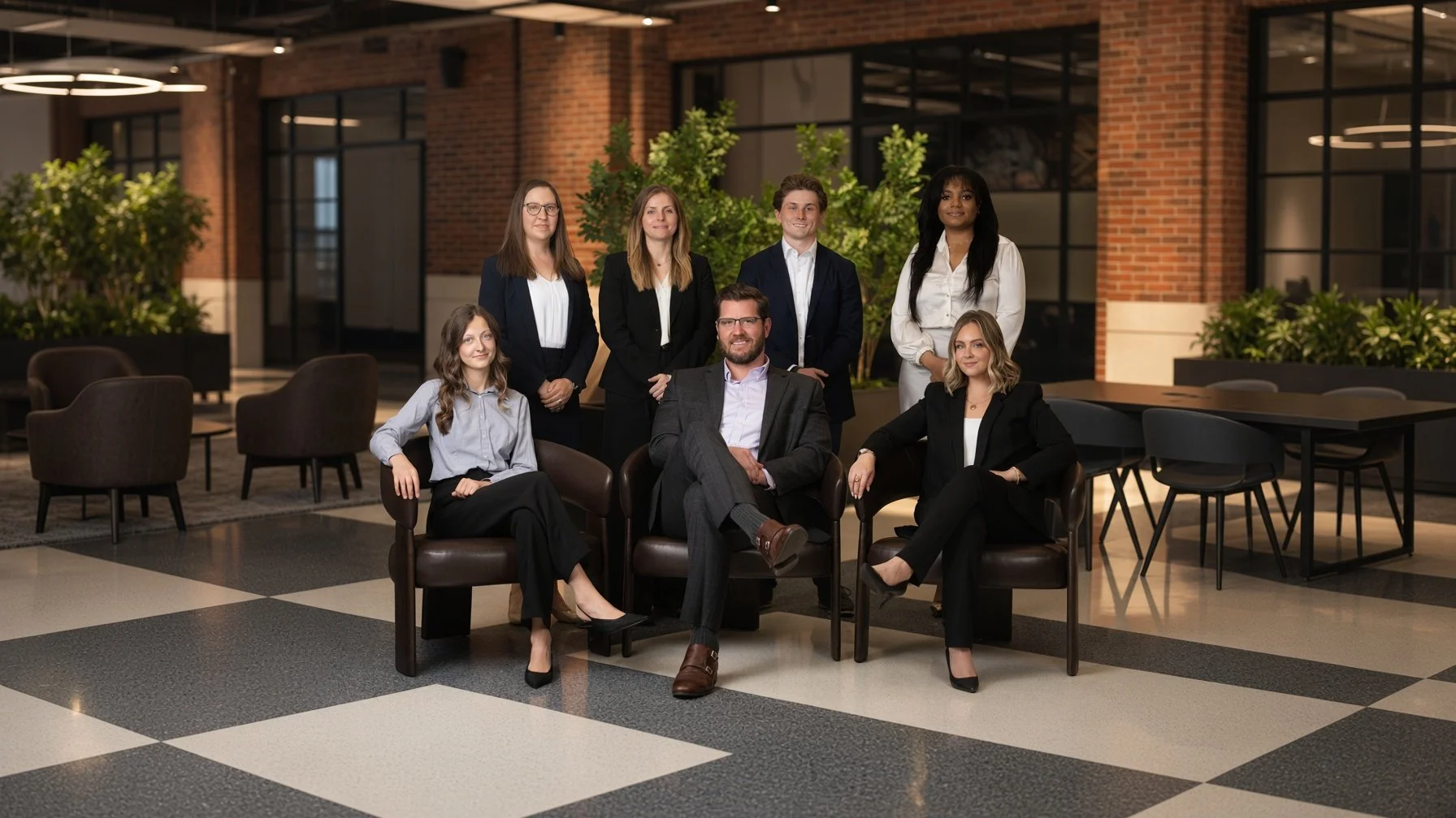 A group of seven diverse professionals, five women and two men, dressed in business attire, posing for a photo in a modern office lobby with brick walls, large windows, and indoor plants.