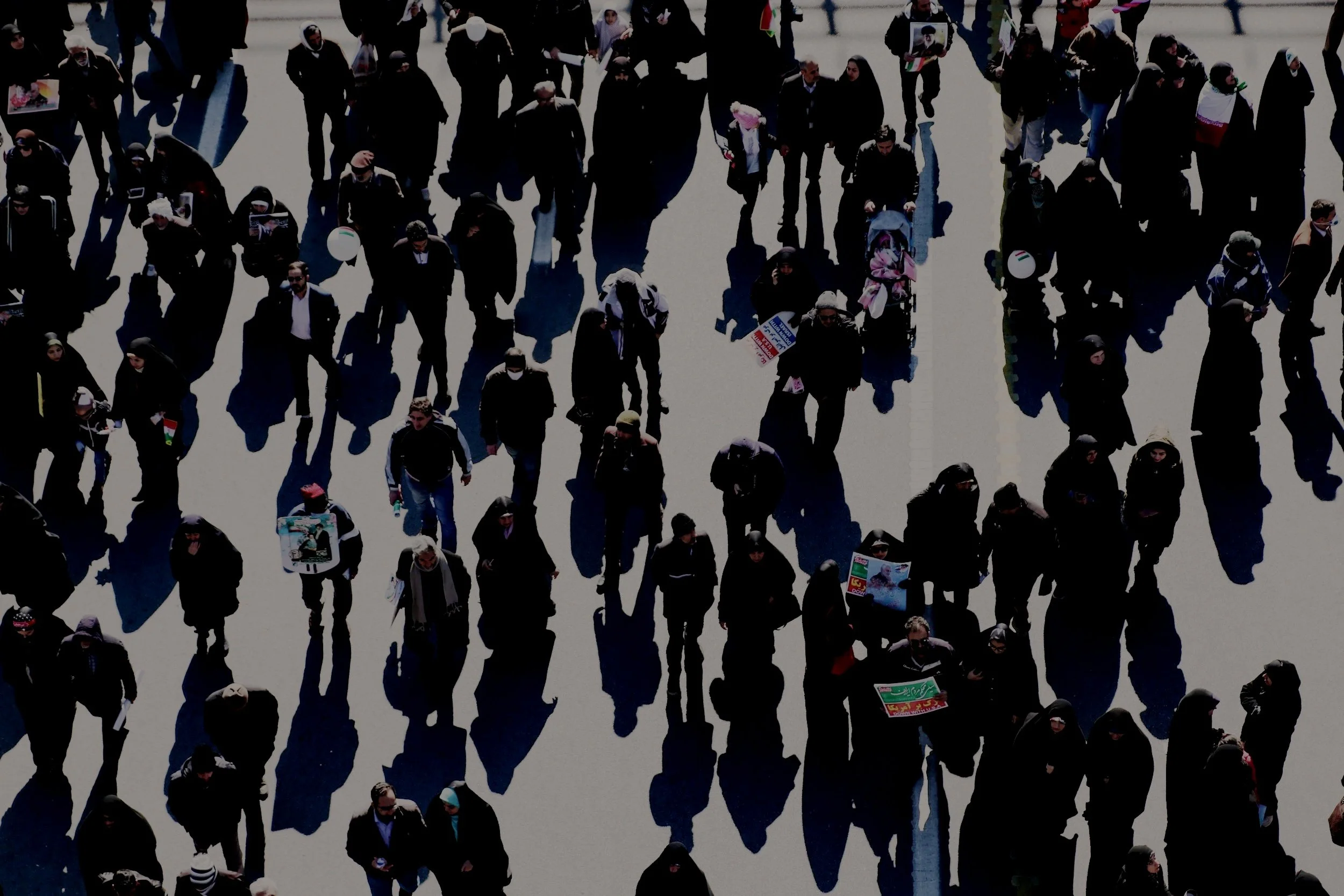 Aerial view of a large crowd of people walking outdoors, some carrying signs and balloons, with shadows on the ground.