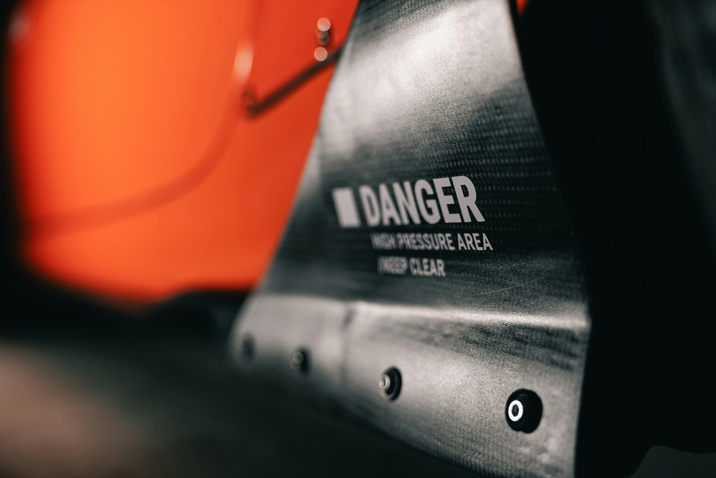 Close-up of a machine with a warning label that reads 'DANGER high pressure area keep clear,' featuring a red and black color scheme with fasteners along the bottom edge.