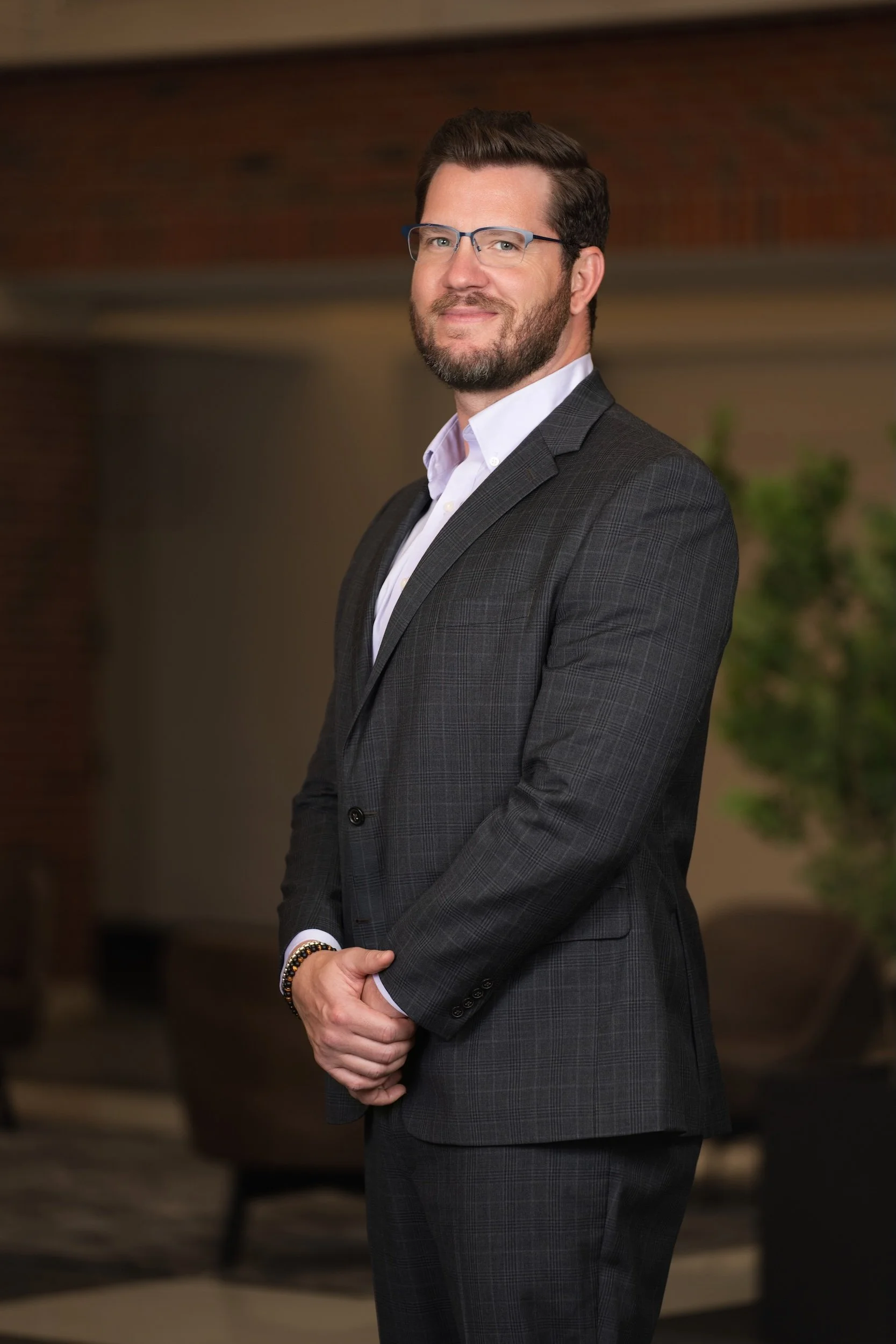A man with glasses and a beard wearing a gray checkered suit and white shirt standing indoors with a blurred background of a brick wall and greenery.