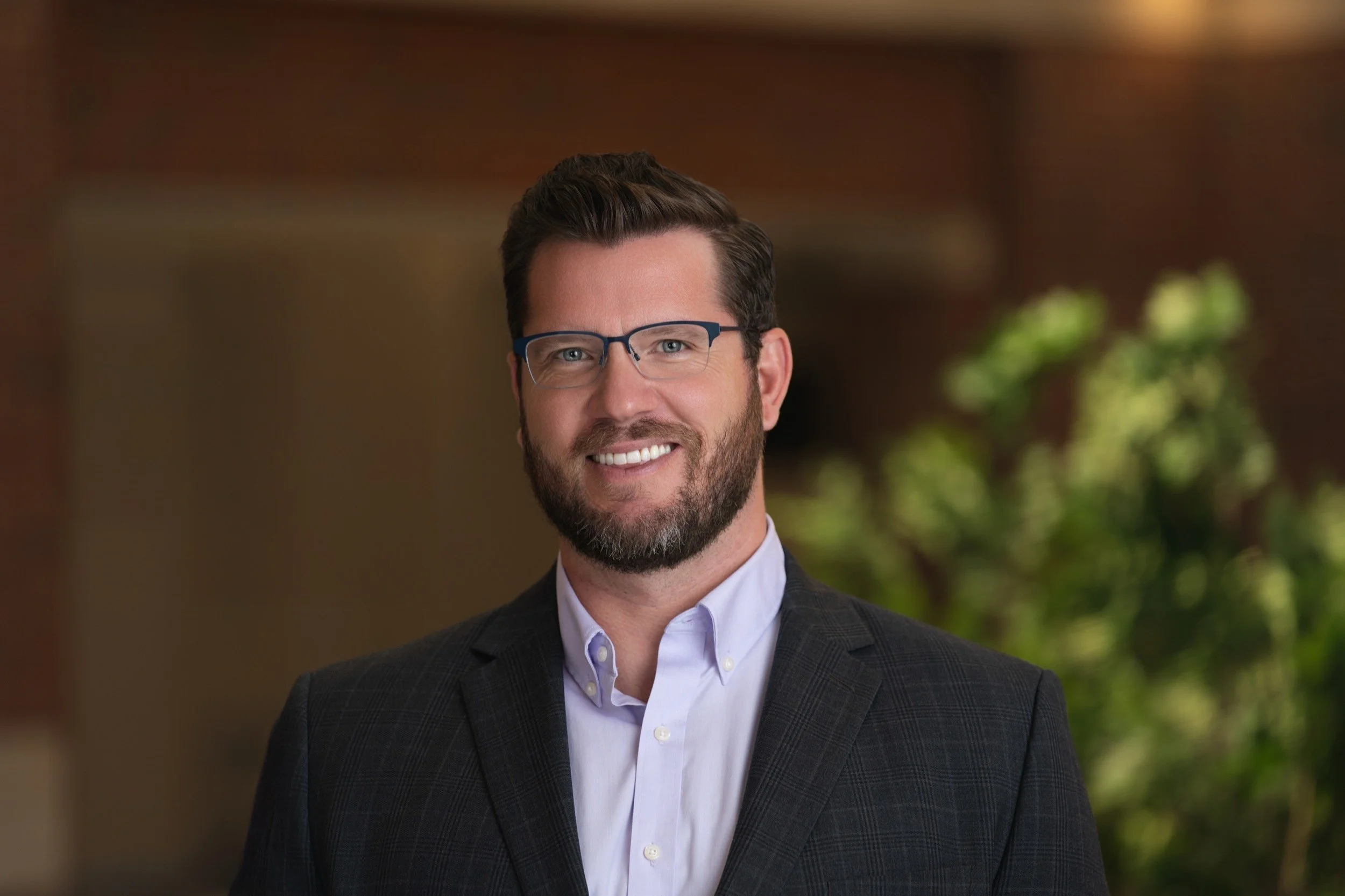 A man with dark hair and a beard wearing glasses, a white shirt, and a dark blazer, smiling in a professional setting with blurred greenery in the background.