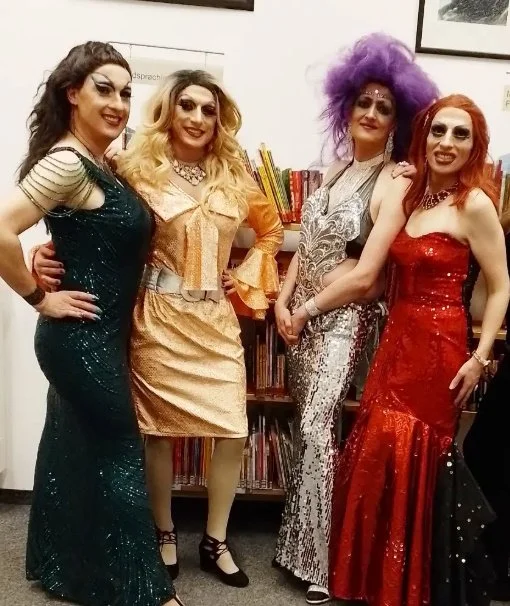 Die KeinKunst Drags Köln - in auffälligen, glamourösen Kostümen und Make-up, stehen nebeneinander in einem Raum mit Büchern im Hintergrund.