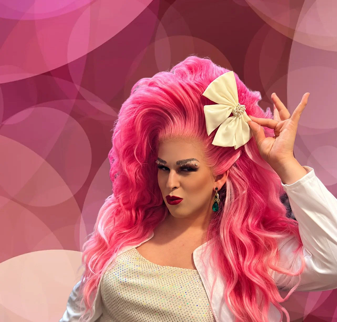 Virginia White mit pinken, gelockten Haaren, auffälligem Make-up, einer großen weißen Schleife im Haar, roten Lippen und grüner Ohrringen posiert vor einem pinken, geometrischen Hintergrund.
