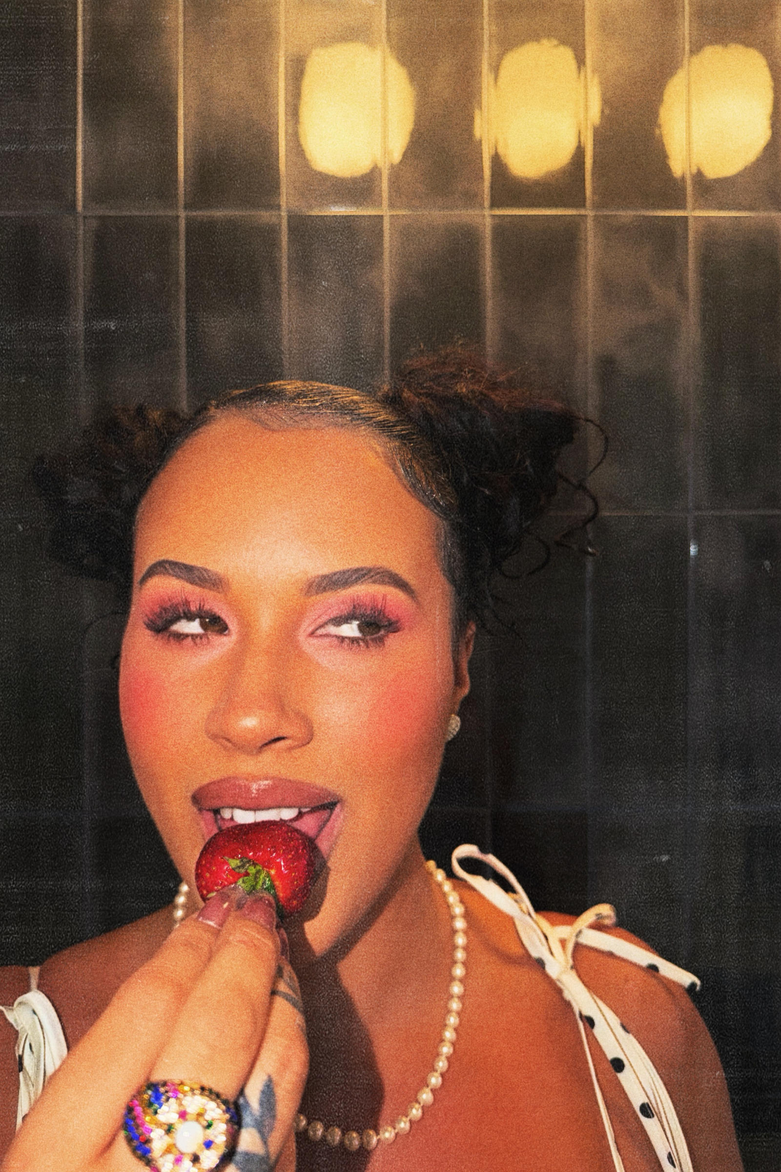 A woman with styled hair and makeup, wearing a pearl necklace and polka dot dress, is about to eat a strawberry.