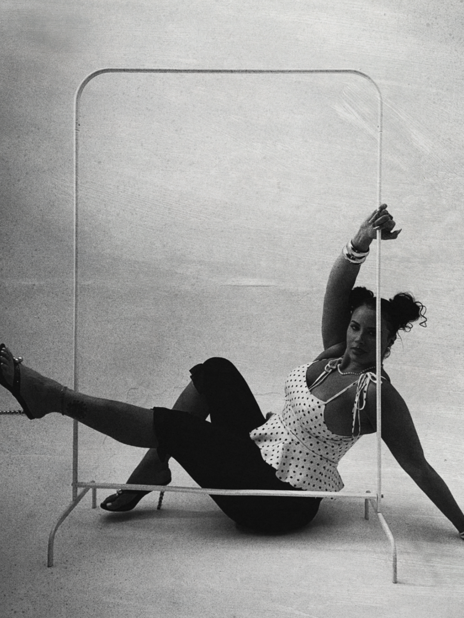 Black and white photograph of a woman lying on a bench in a dance studio, holding a vertical pole, wearing a polka dot blouse, black pants, and high heels, with her hair in a high bun.