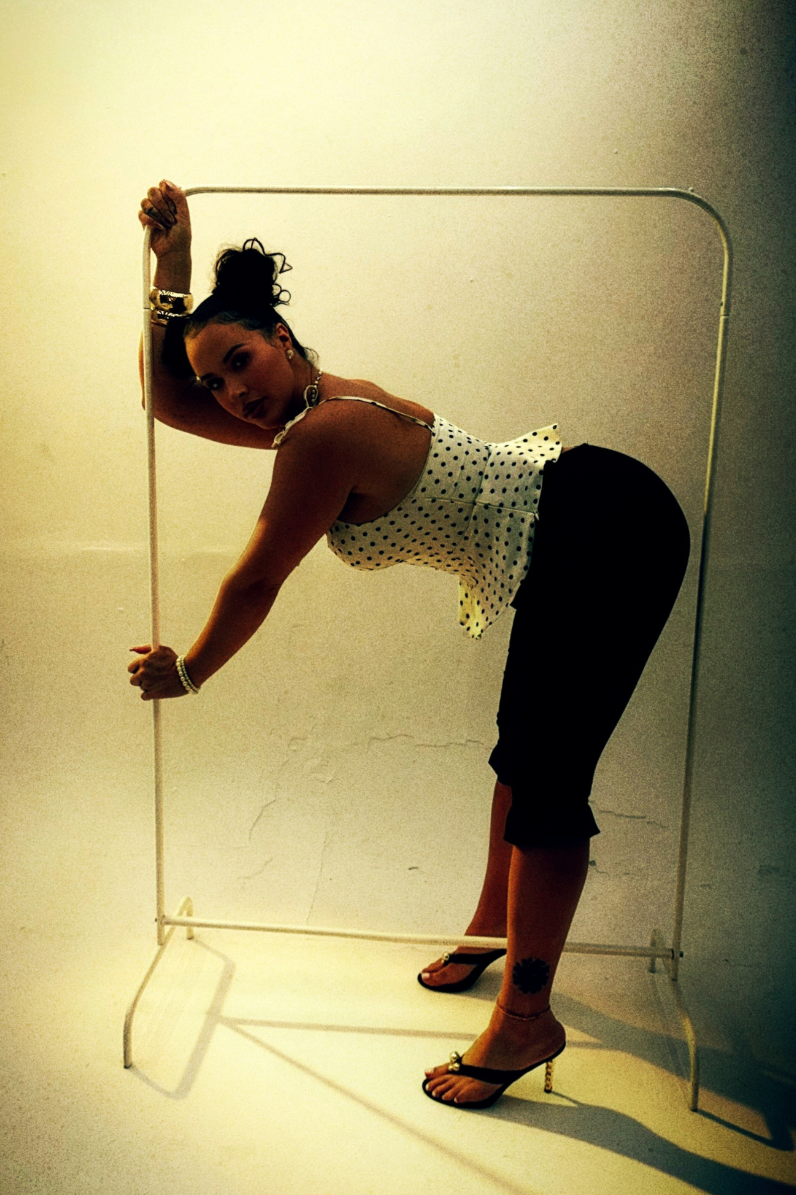 A woman with dark hair in a bun, wearing a polka dot top and black pants, poses against a beige wall with a metal frame, wearing high heels with gold heels and jewelry.