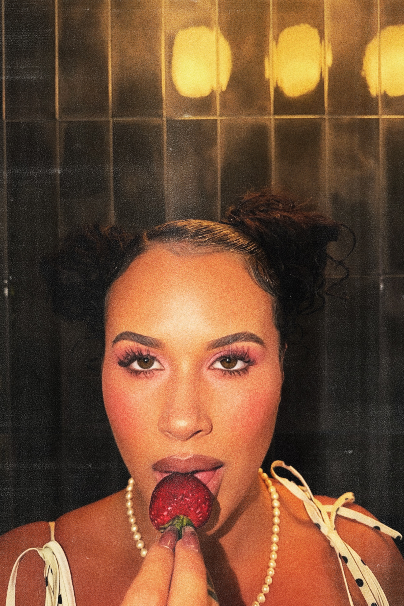 Close-up of a woman with dark, curly hair styled in two buns, holding a strawberry near her lips, wearing pearl earrings, makeup with pink eyeshadow and bold eyebrows, against a black tiled background with warm yellow lights.