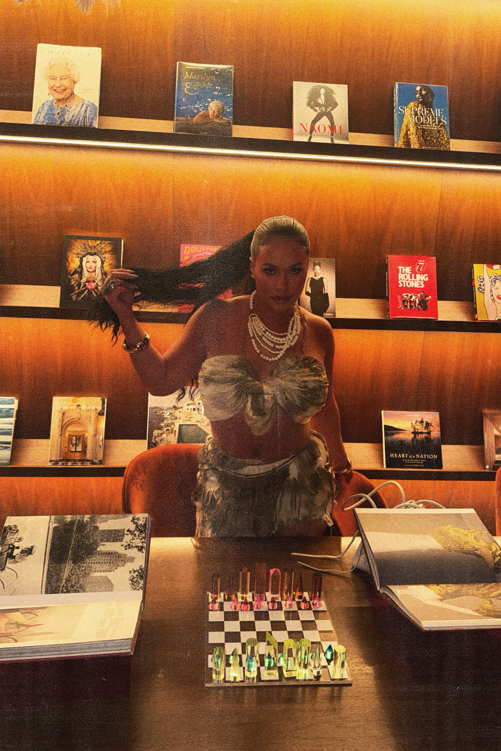 A woman dressed in an elegant outfit with pearl jewelry and a large bow on her top, sitting at a table in a bookstore or library, surrounded by books and displayed chess pieces, with shelves of books in the background.