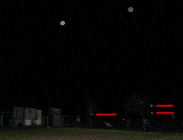 Dark night scene with two bright celestial objects in the sky and some houses or buildings with red lights in the foreground.