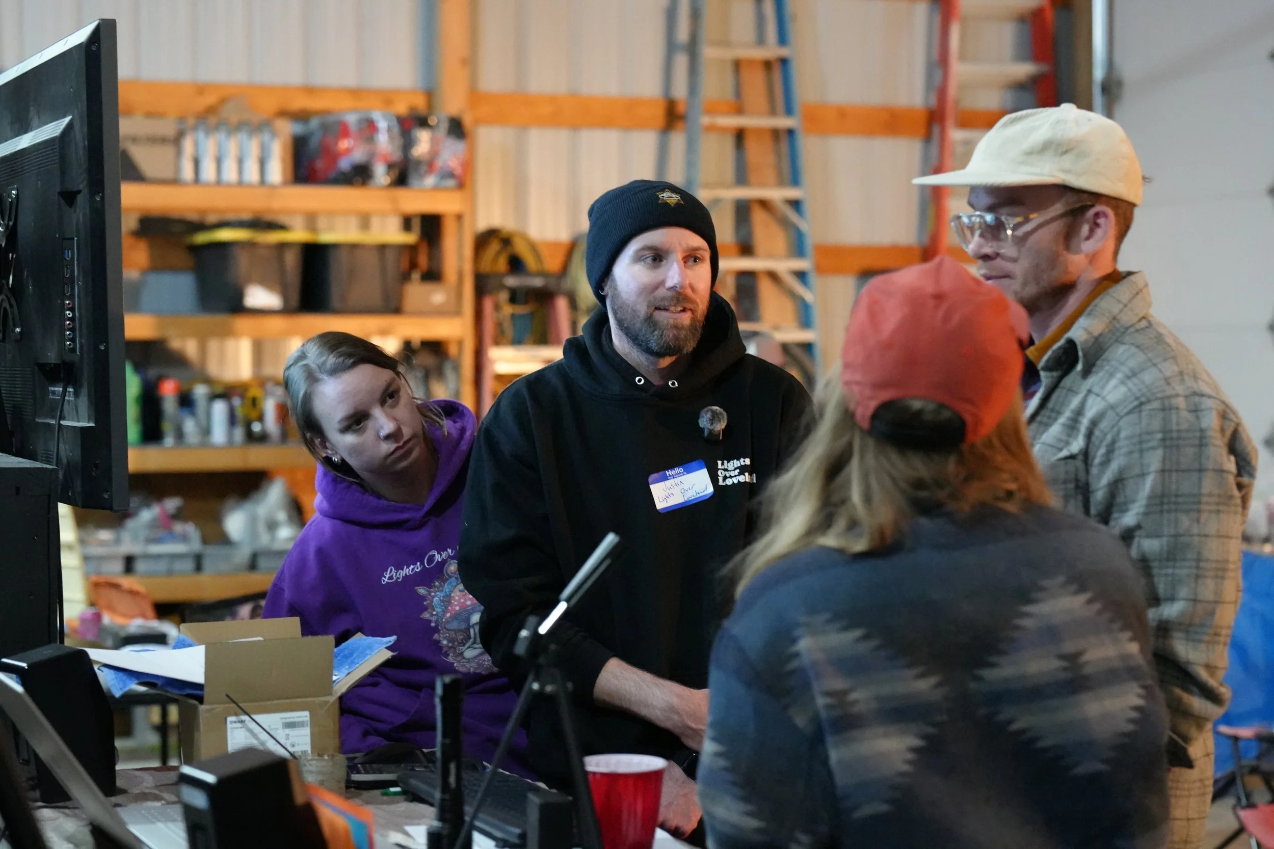 Several people gather around a computer in a workshop space, with shelves holding various items like helmets and boxes in the background.