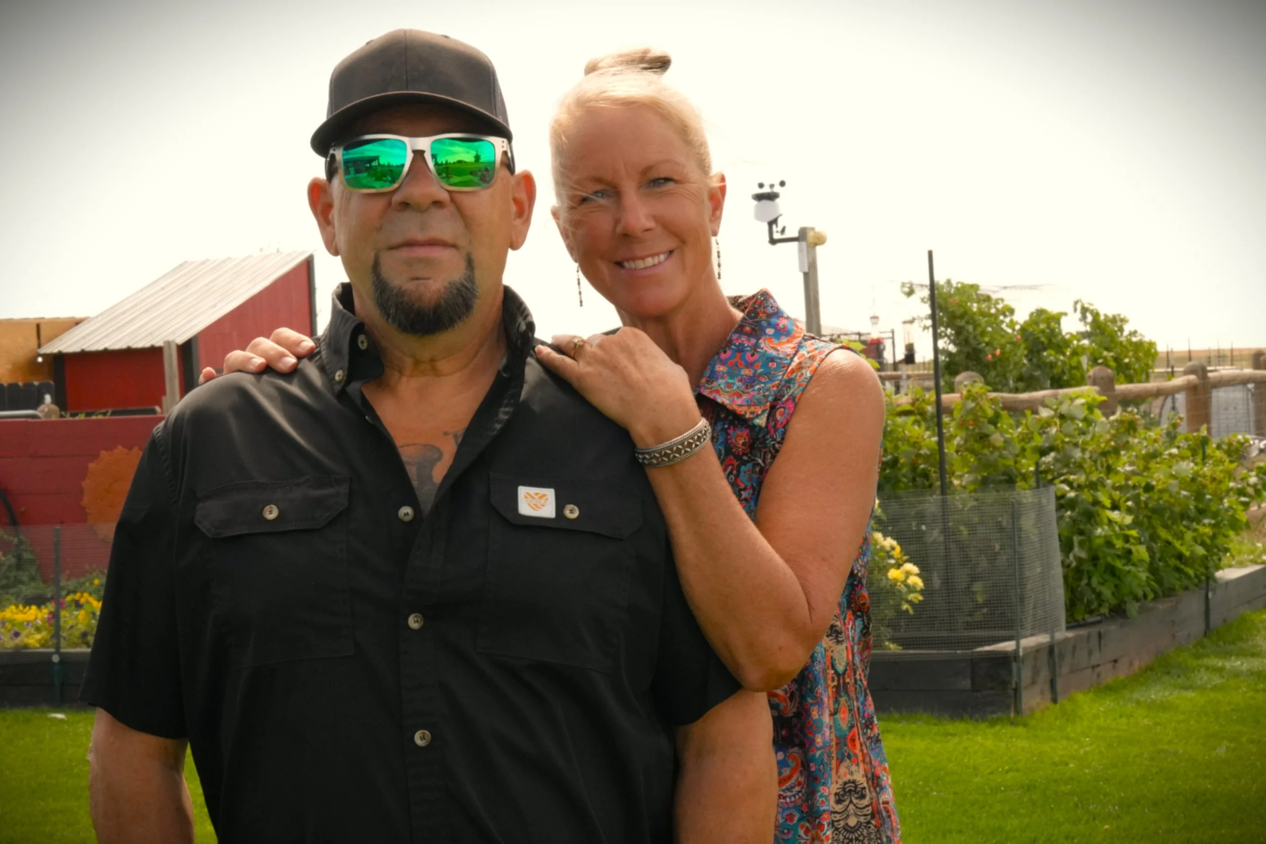 A man wearing sunglasses, a black cap, and a black shirt standing next to a woman with blonde hair in a patterned dress. They are outdoors in a garden or farm area with plants, flowers, a red barn, and a weather vane in the background.