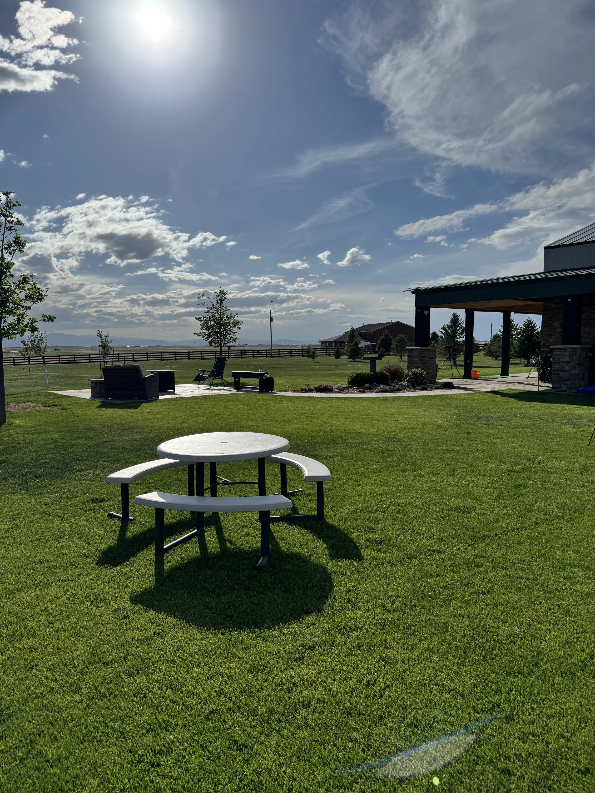 A backyard scene with a green lawn, outdoor seating, a small round white table, a building with a porch, trees, and a partly cloudy sky with the sun shining brightly.