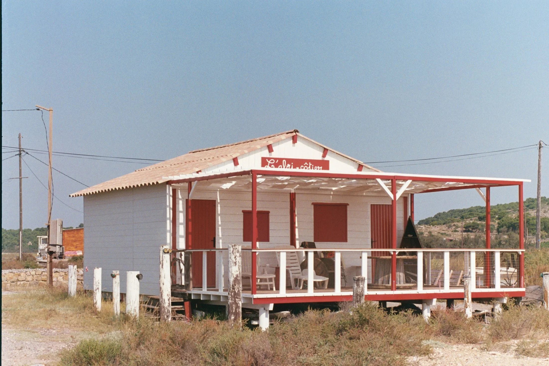 Une petite maison en bois blanc avec un toit en tuiles rouges, située dans un environnement rural avec un terrain en herbe et des poteaux électriques à proximité.