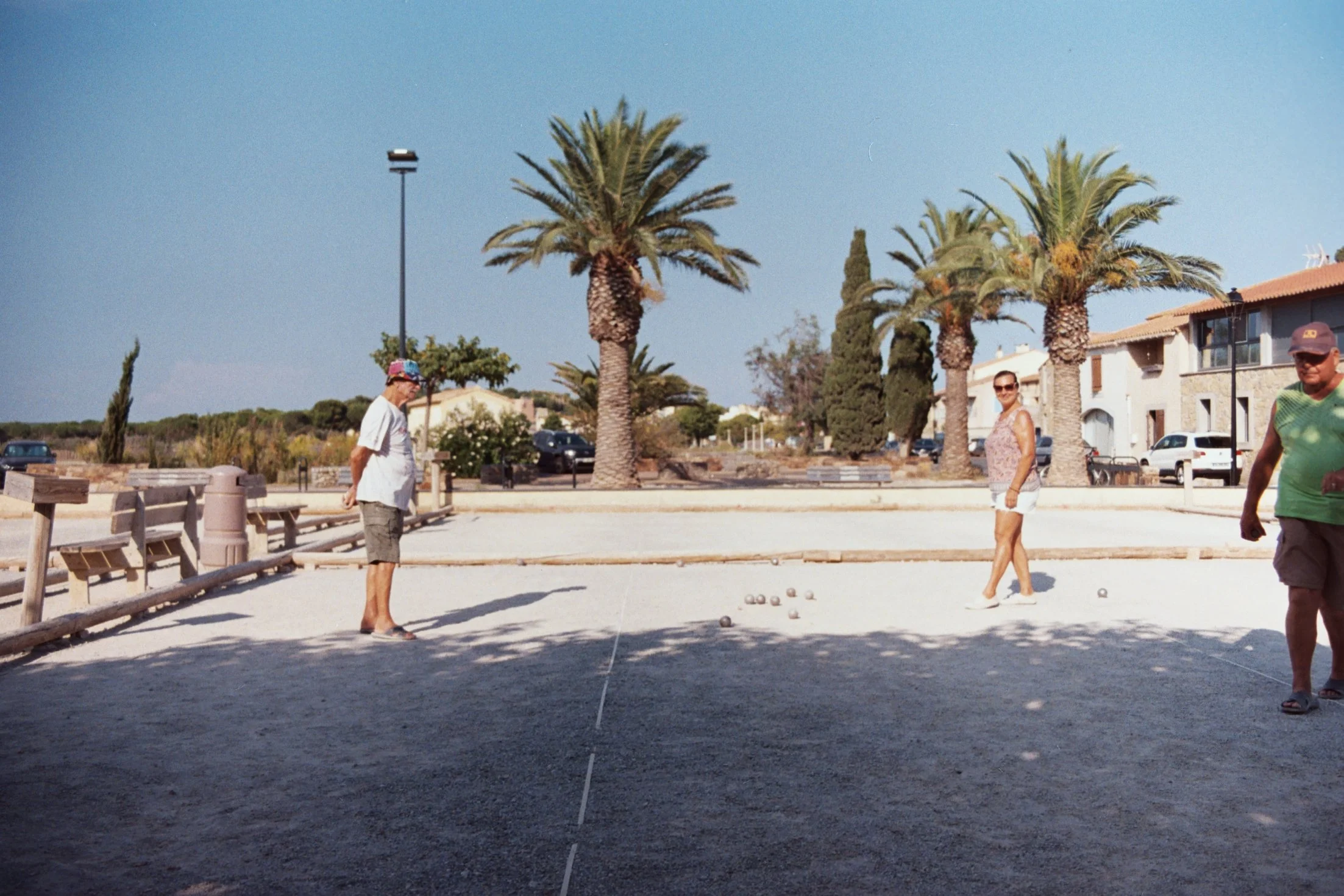 Trois personnes jouent à la pétanque dans un parc ensoleillé, avec des palmiers et des maisons en arrière-plan.