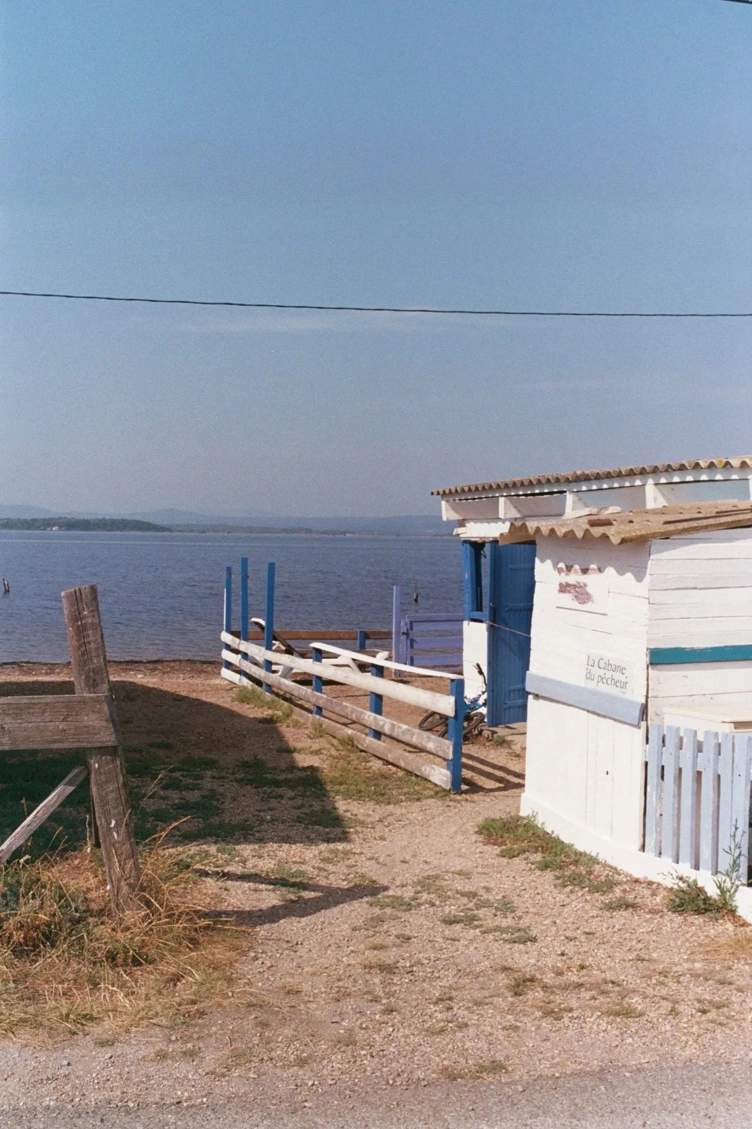 Vue de côté d'une petite structure en bois blanc avec un toit en tuiles, surplombant un fence en bois peint en blanc et bleu, avec la mer en arrière-plan et un ciel en journée claire.
