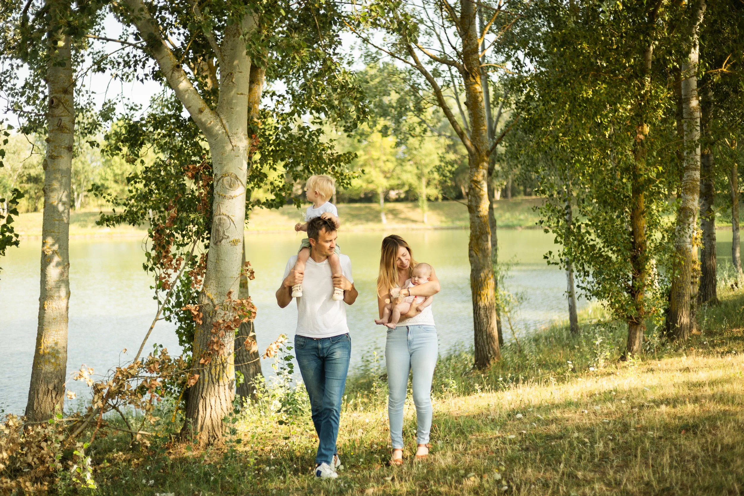 Un groupe de quatre personnes, deux adultes et deux enfants, se promènent dans un parc près d'un lac en portant des vêtements décontractés et souriant. Il y a plusieurs arbres autour d'eux et la scène est en plein air en journée.