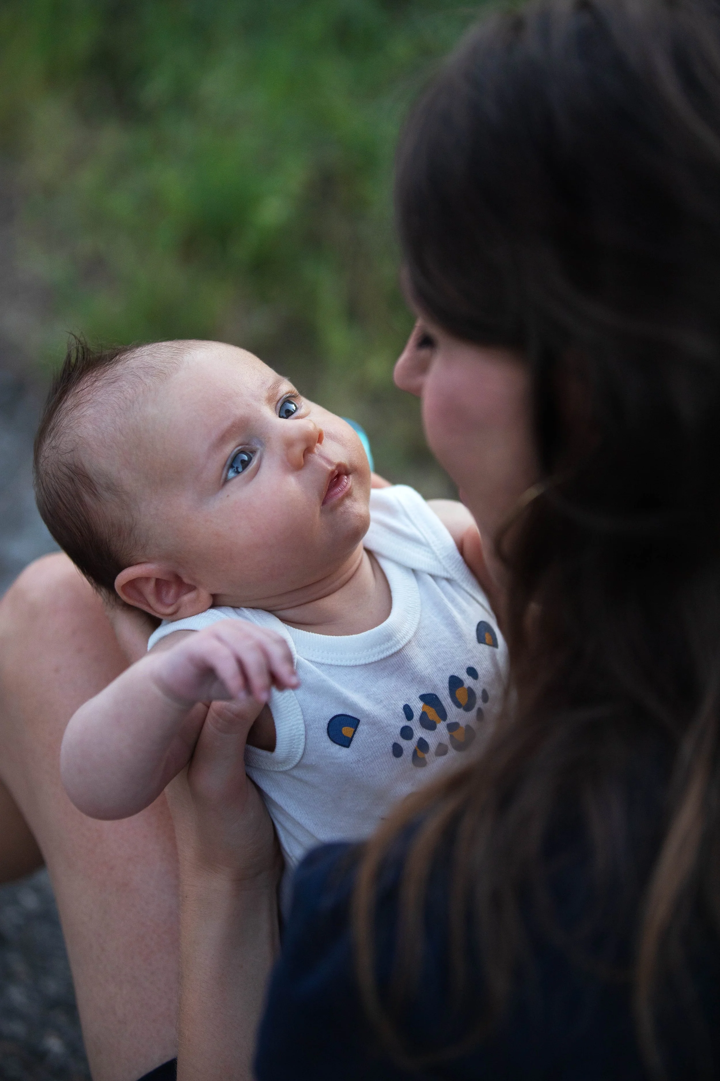 Un bébé regarde une femme dont seule une partie du visage est visible, en arrière-plan un espace dehors avec des végétaux verdoyants.