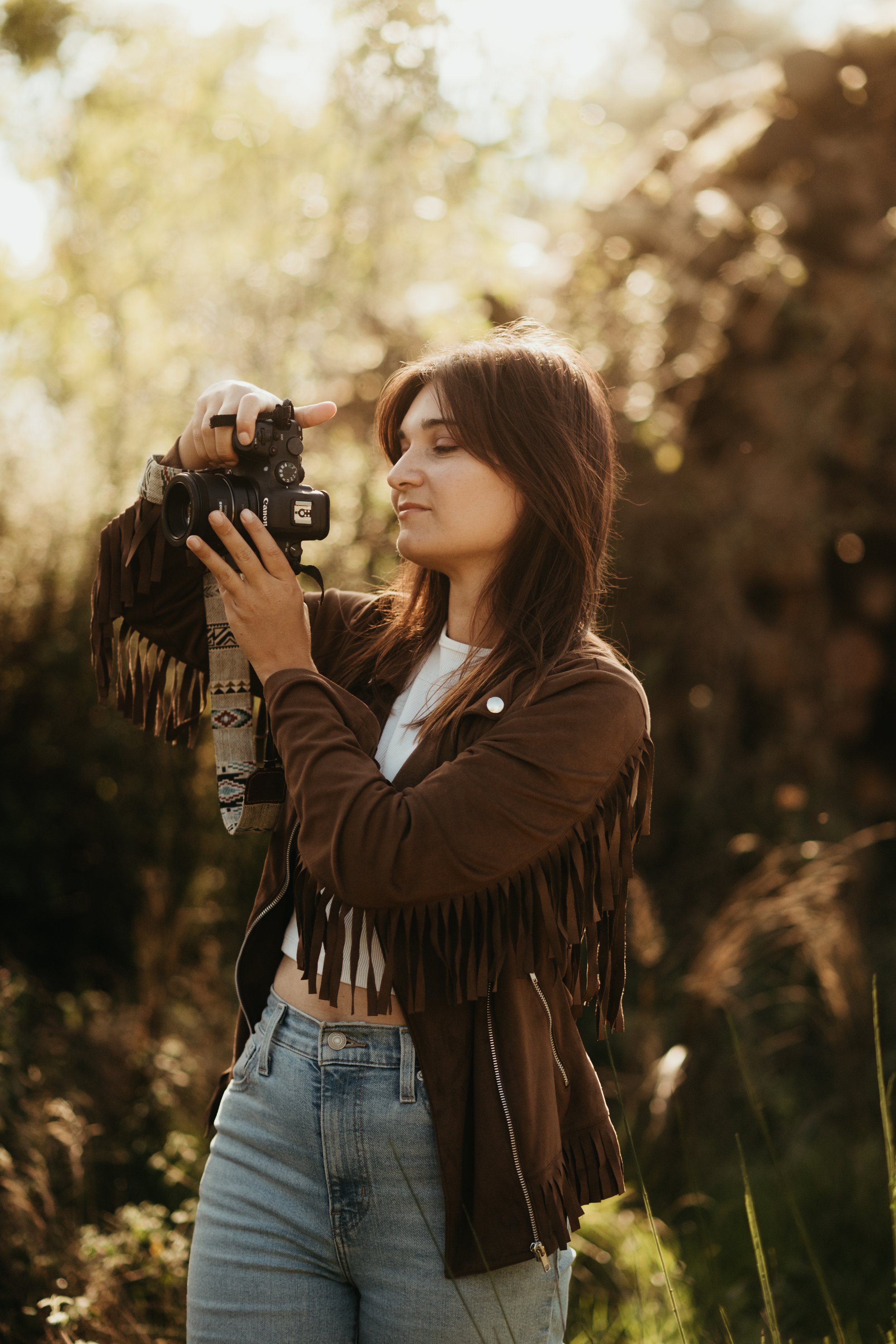 Une femme avec des cheveux longs bruns, portant une veste en cuir marron avec des franges, une chemise blanche, et un jean bleu, tient un appareil photo dans ses mains, en extérieur lors d'une journée ensoleillée.