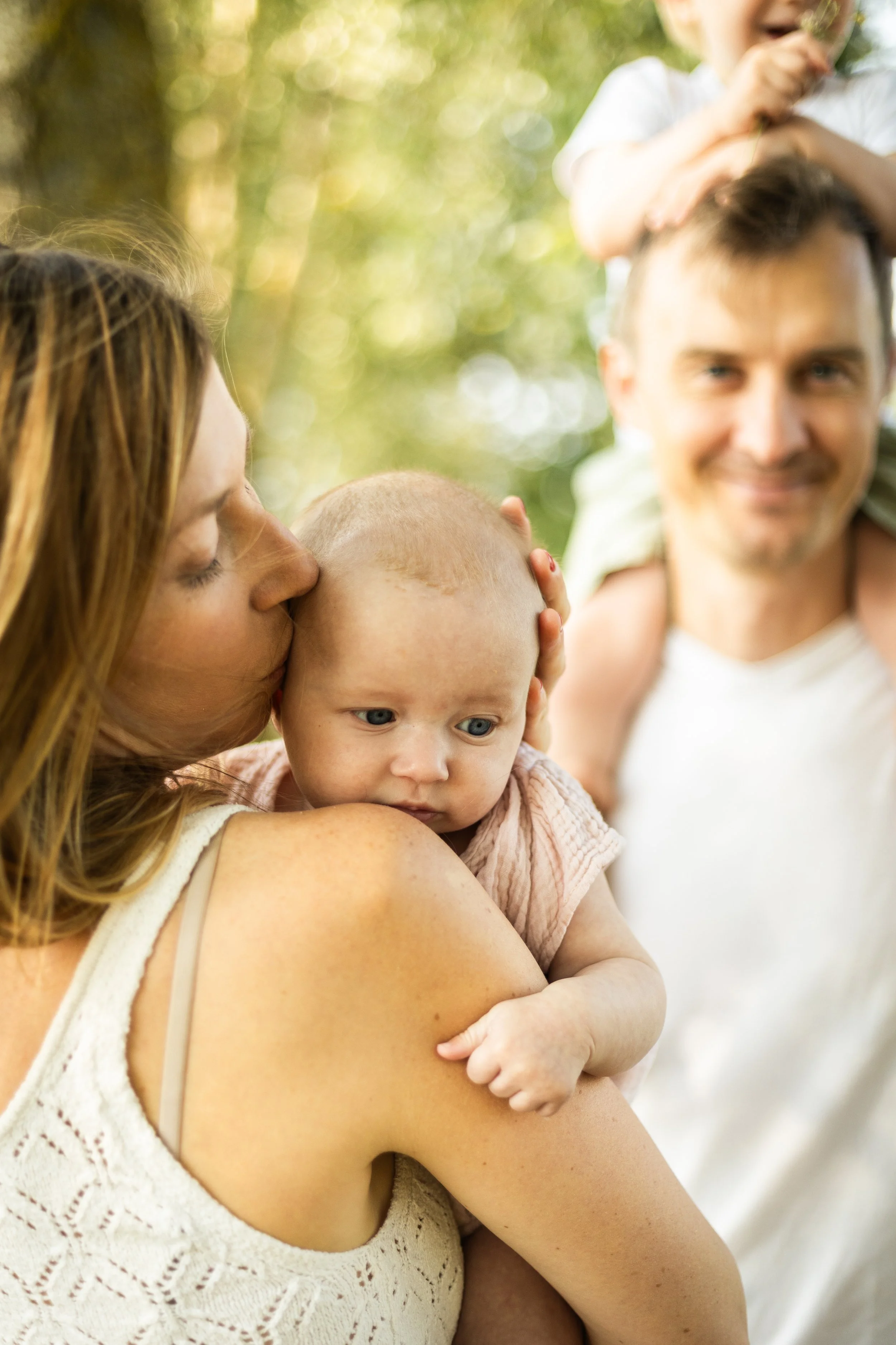 Une famille en plein air, une femme embrasse un bébé sur l'épaule, un homme porte un enfant sur ses épaules, avec des arbres en arrière-plan.