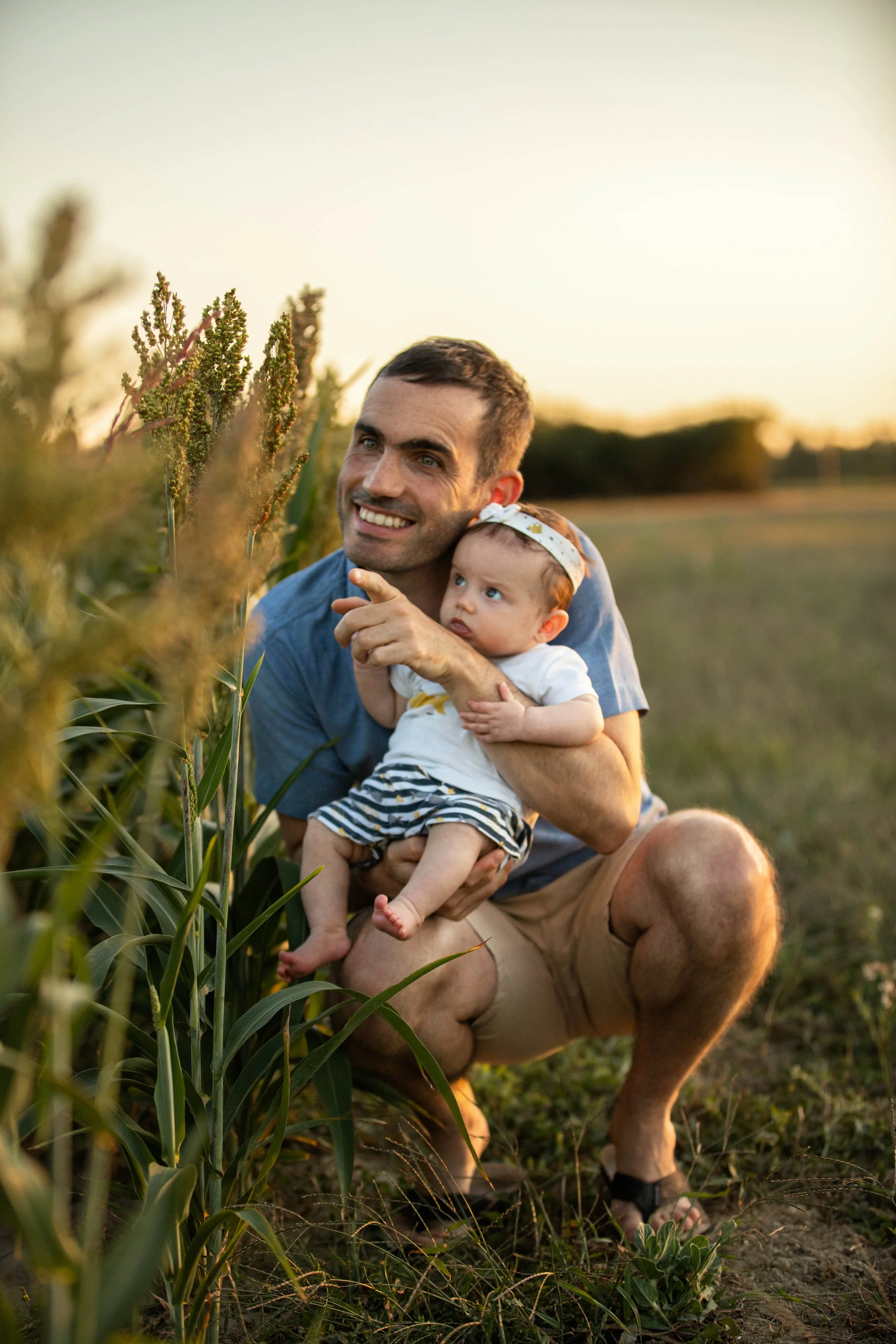 Un homme et un bébé assis dans un champ de cultures, le soleil couchant en arrière-plan, l'homme pointant du doigt quelque chose, tous deux souriants.