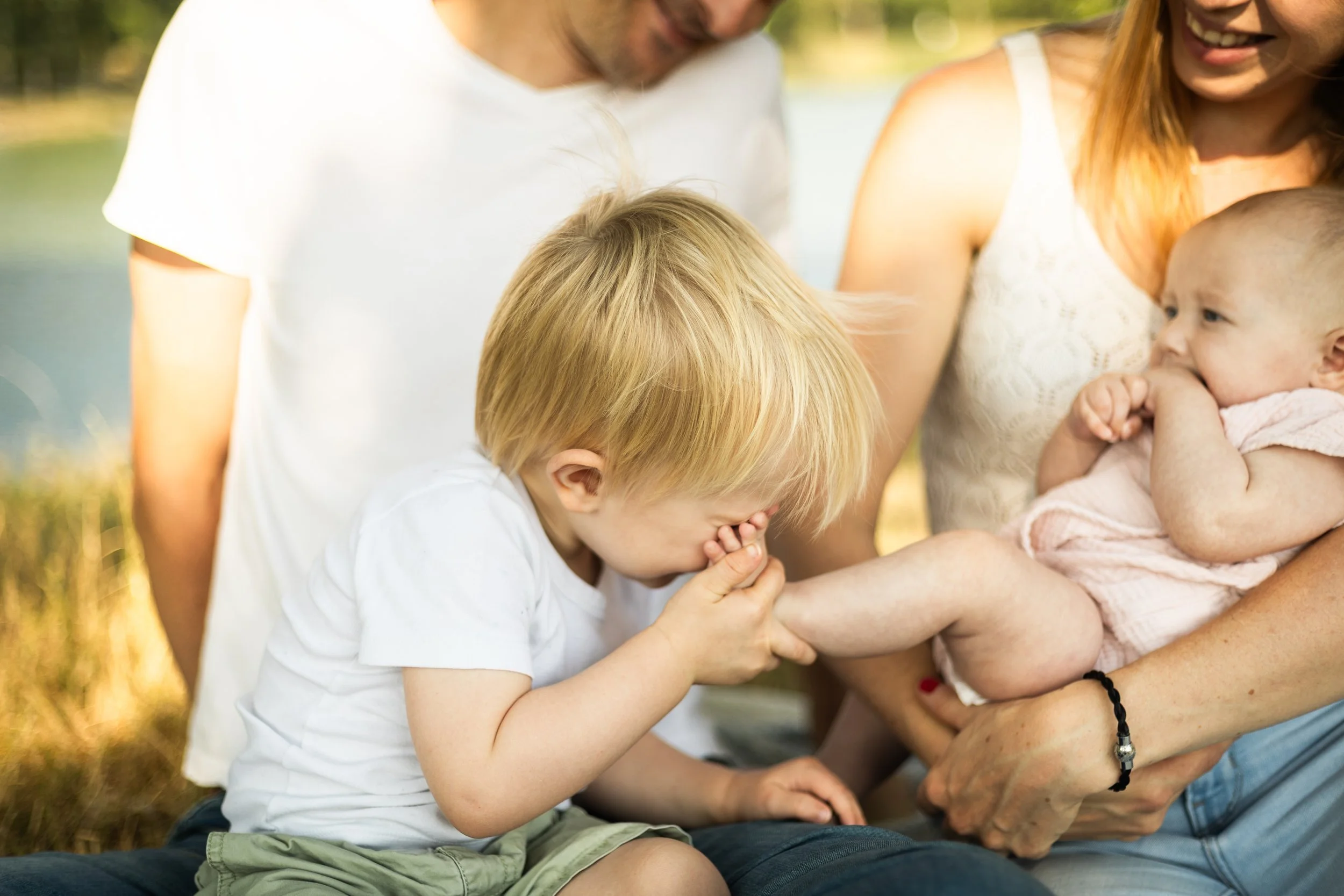 Une famille de quatre personnes, deux adultes et deux enfants, partage un moment de bonheur en plein air près de l'eau. La mère tient un bébé, tandis qu'un garçon prière ou prie avec ses mains jointes, tous souriant et se trouvant sur une surface herbeuse ou près de l'eau.