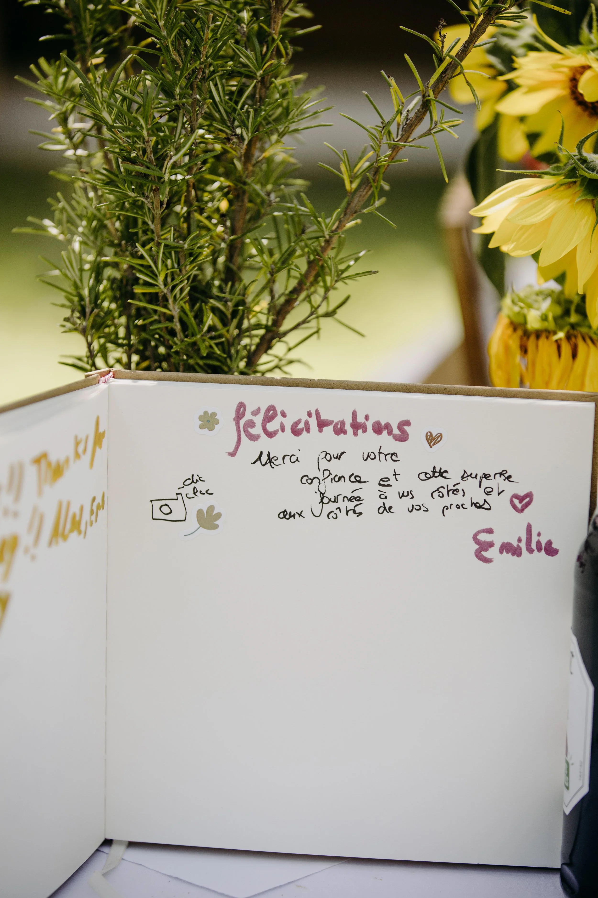Une carte de remerciement écrite à la main avec un motif floral, placée devant un bouquet de fleurs jaunes et un buisson vert. La carte exprime des remerciements pour la confiance et une journée spéciale.