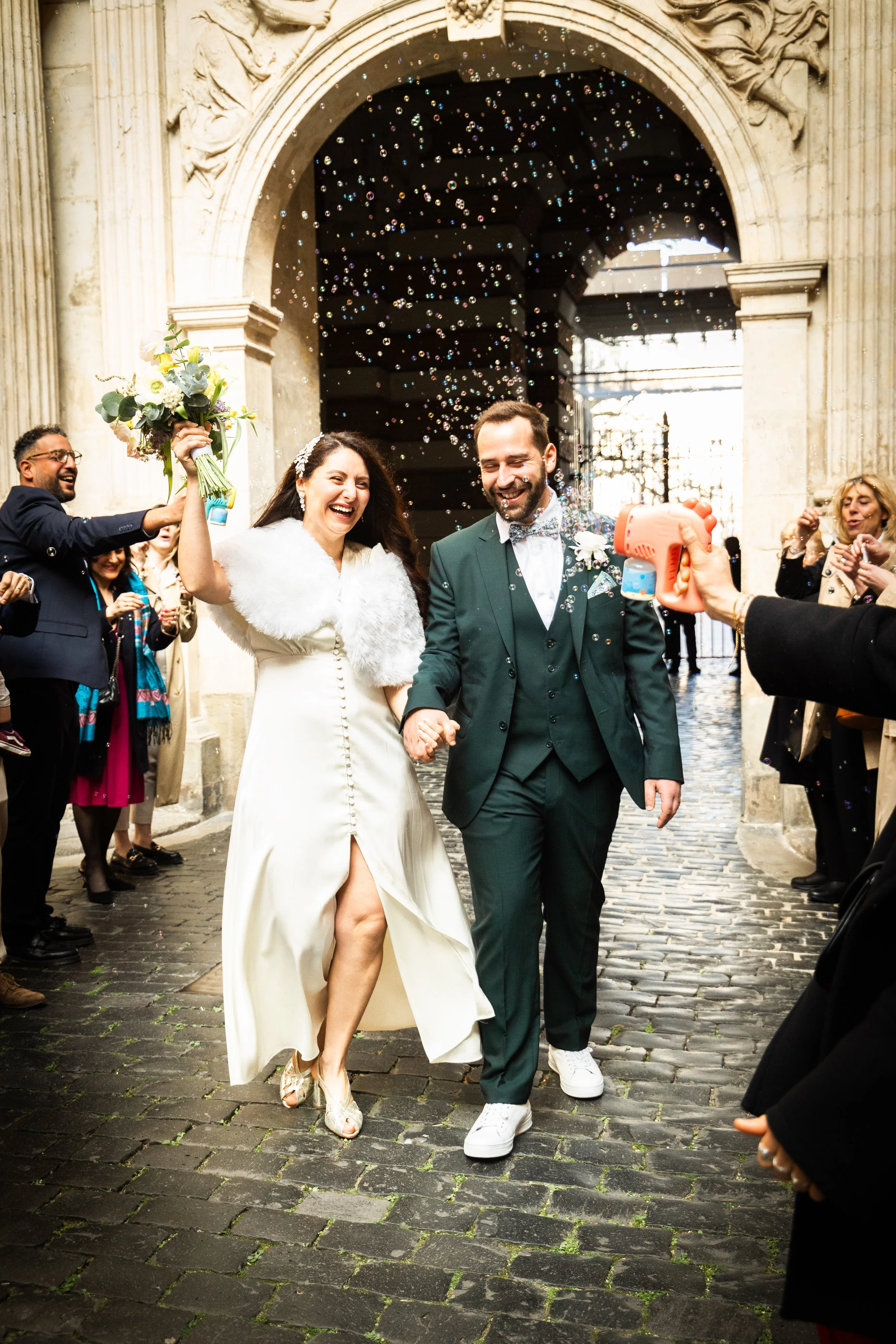 Un couple de mariés souriants en train de marcher main dans la main lors de leur mariage, entourés d'invités célébrant sous une arche en pierre.