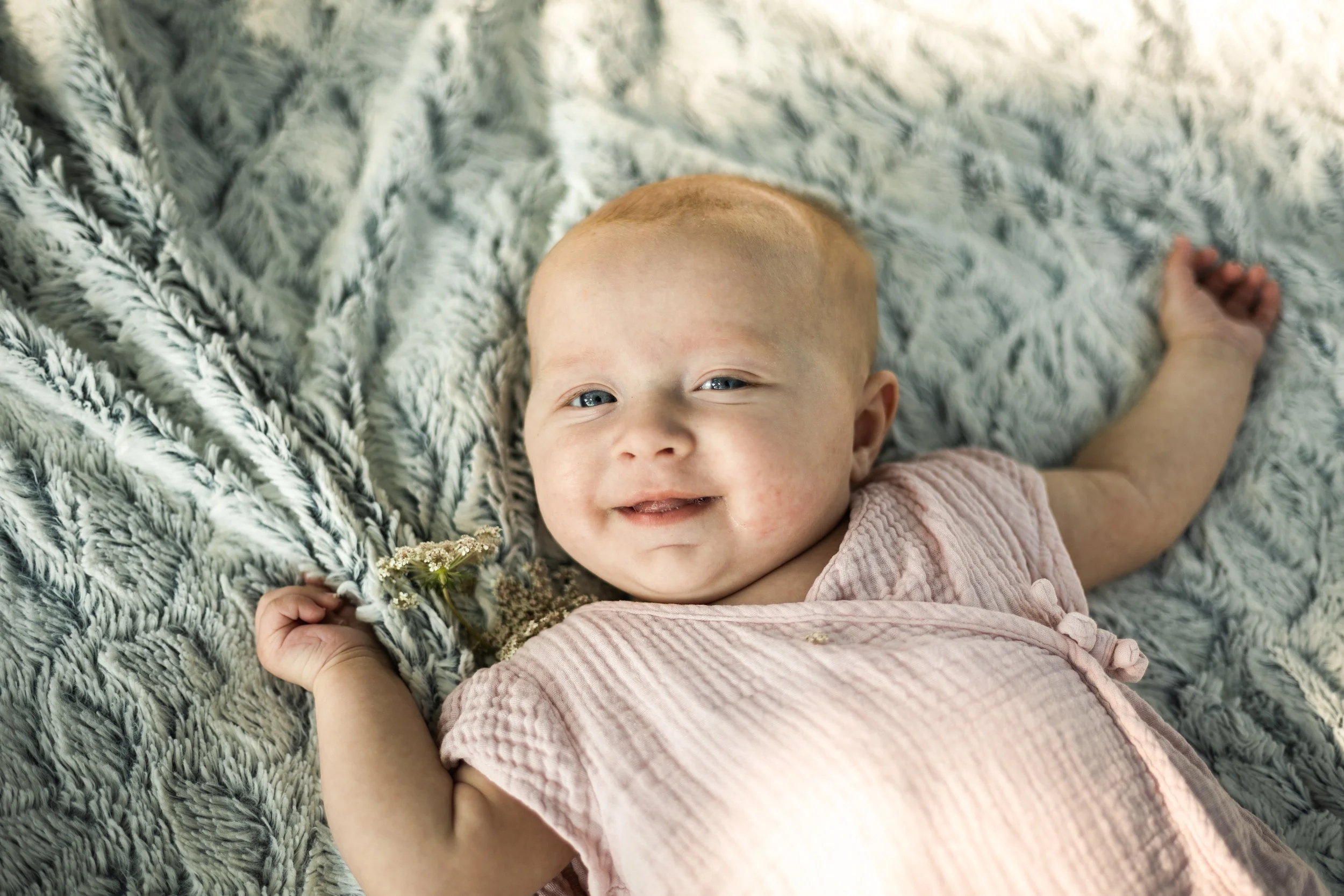 Un bébé avec des cheveux roux, allongé sur un lit recouvert d'une couverture en fausse fourrure grise et blanche, tenant une petite fleur dans la main.