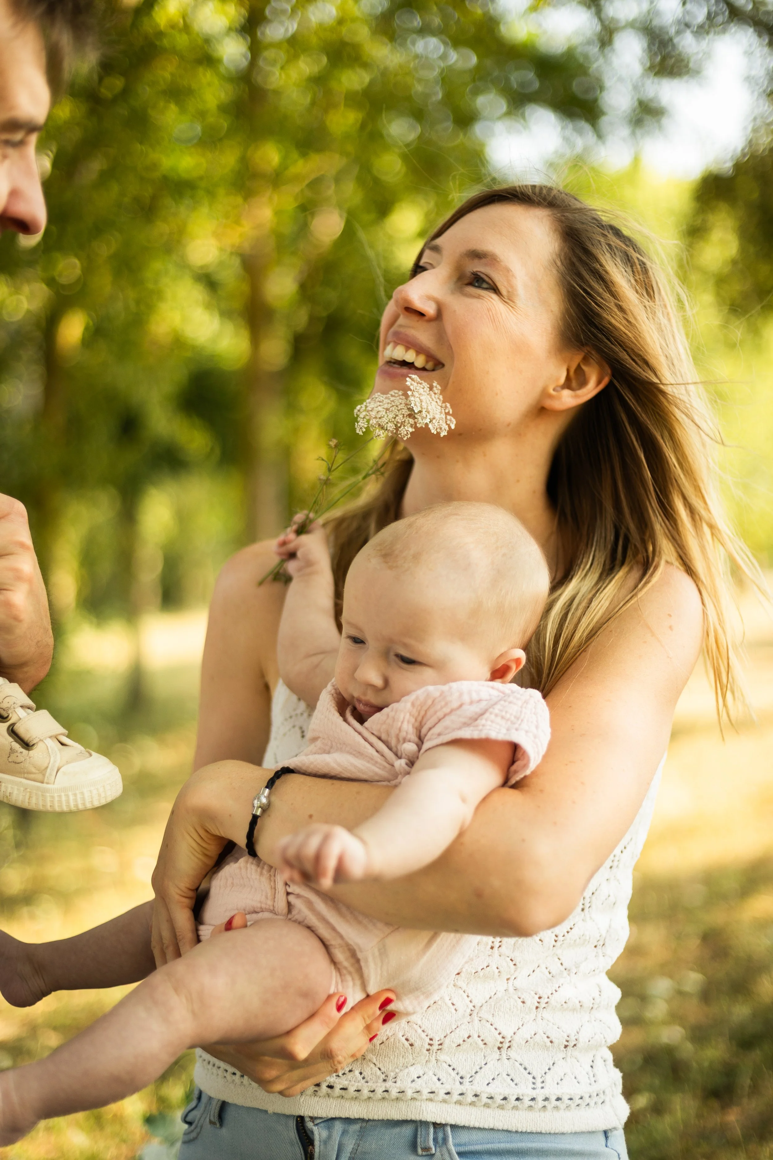 Une femme souriante tient un bébé dans ses bras dans un parc ensoleillé, pendant que quelqu'un lui tend une chaussure adidas.