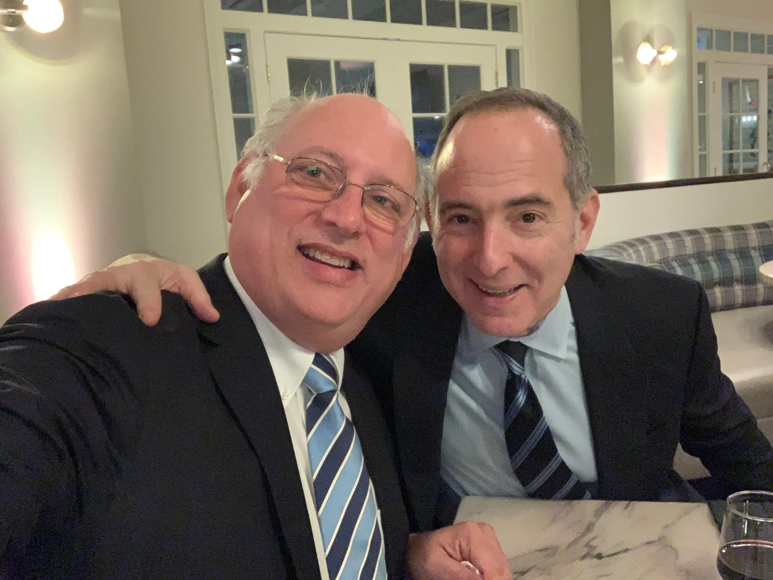 Two men dressed in suits enjoying a meal together, smiling and posing for the photo inside a well-lit room with large windows and wall-mounted lights.