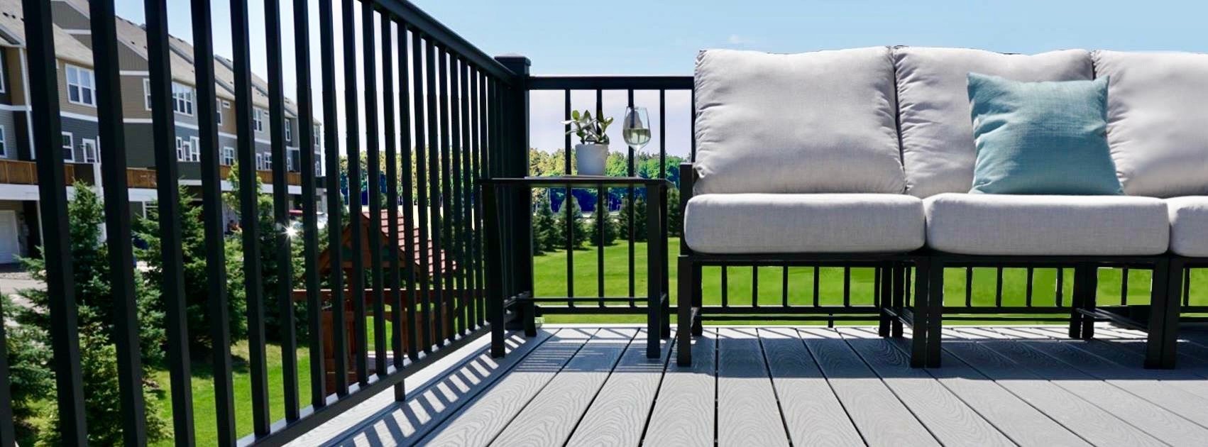 View of a balcony with a black railing, a white outdoor sofa with a blue pillow, a small side table with a plant and a glass of water, overlooking a green lawn and trees, with apartment buildings in the background.