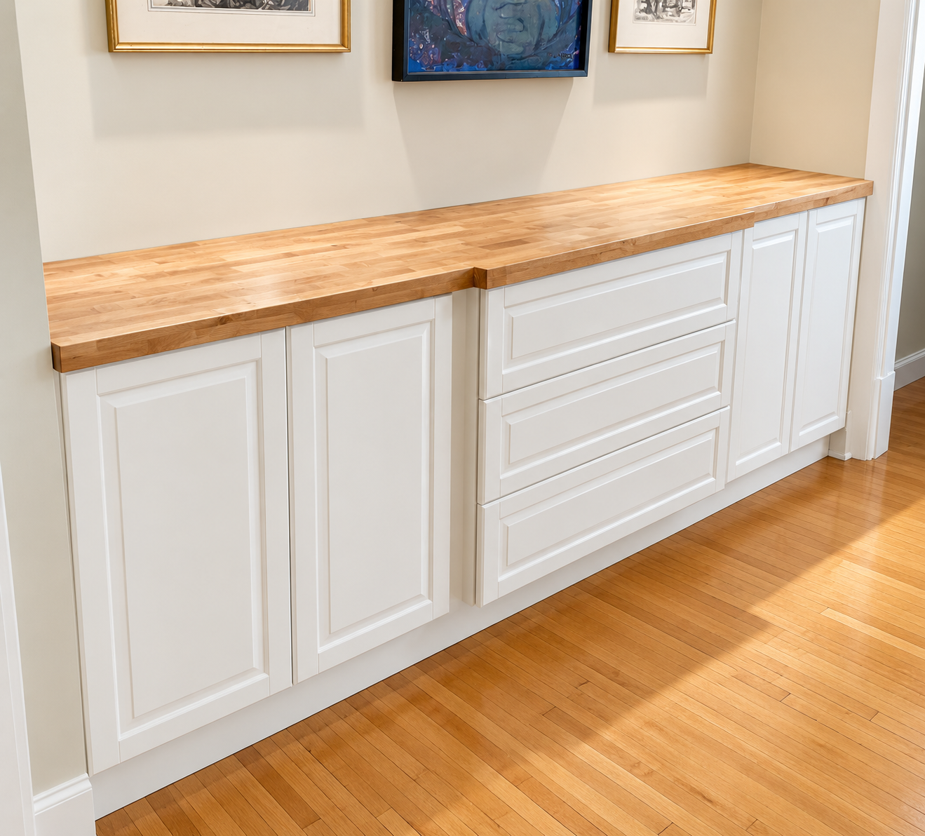 A kitchen or dining area cabinet with a wooden countertop and white paneled cabinet doors, with artwork hanging on the cream-colored wall above the cabinet.