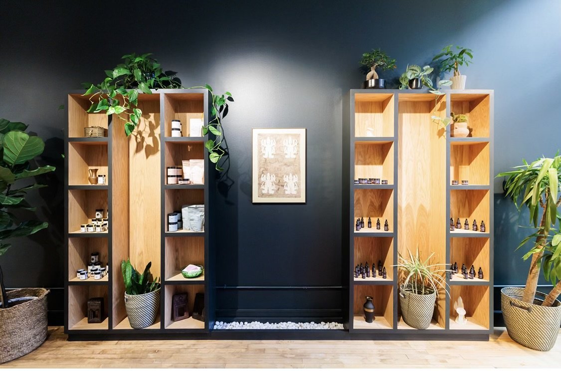 Interior of a store with two wooden shelves against a dark wall, displaying bottles and products, with potted plants on both sides and a framed picture on the wall.