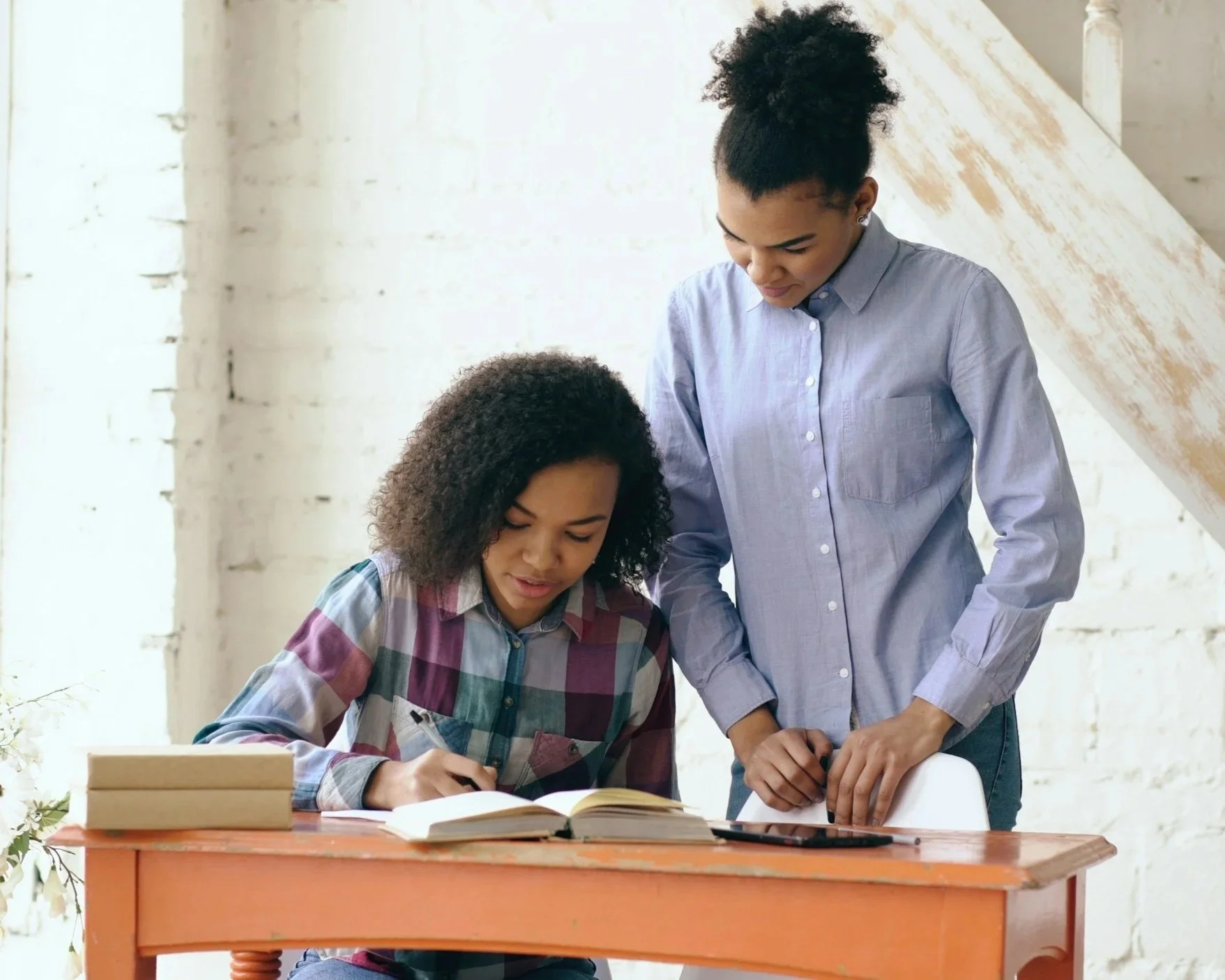 Two women working on a project together at a table in a bright, rustic room. One woman is sitting with an open book, writing, and the other woman is standing, leaning over to look at the book.