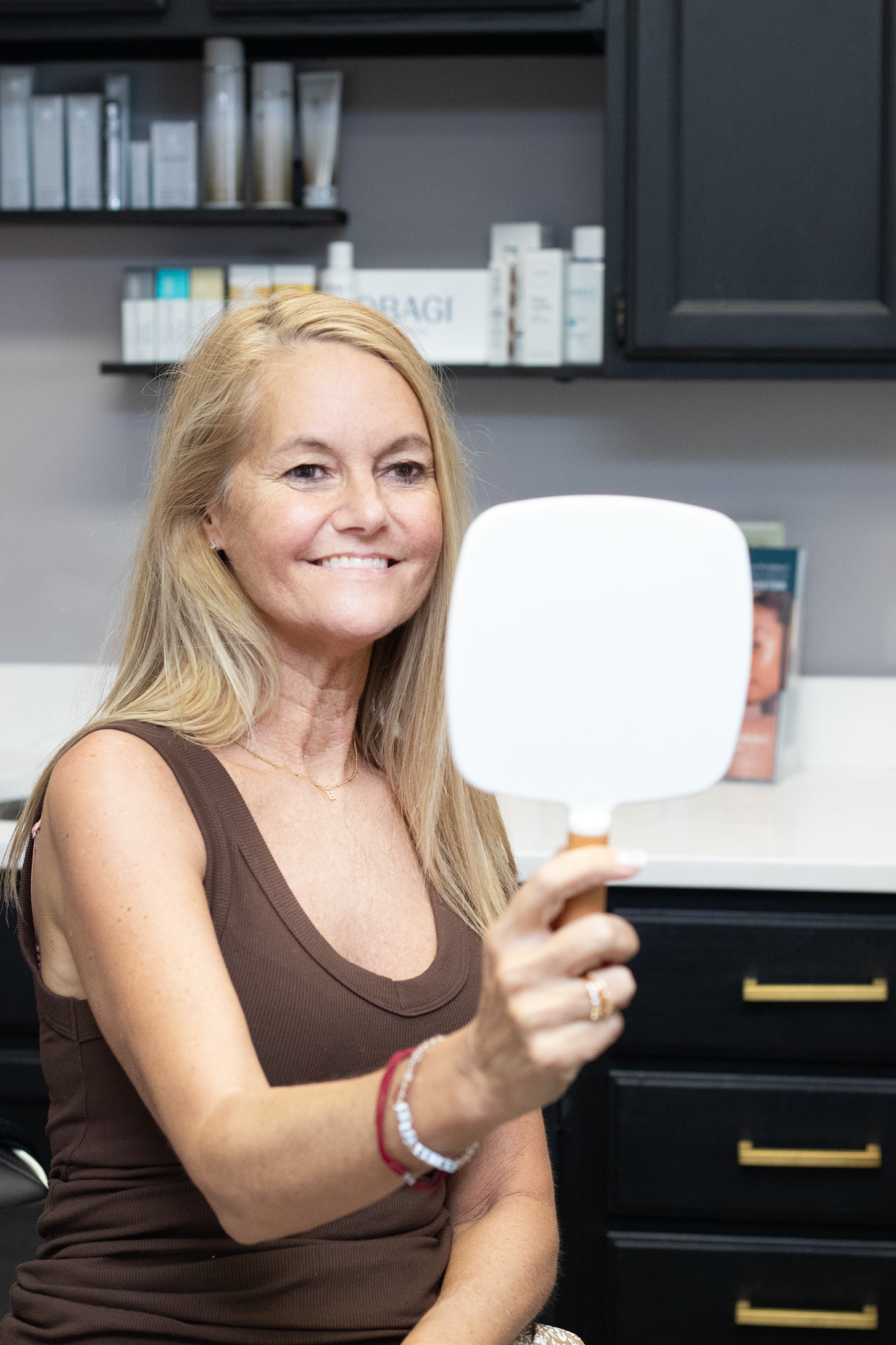 A smiling woman with blonde hair wearing a brown sleeveless top, holding a mirror in her right hand, with shelves of skincare products in the background at Rejuvenate Wellness in Florence Kentucky