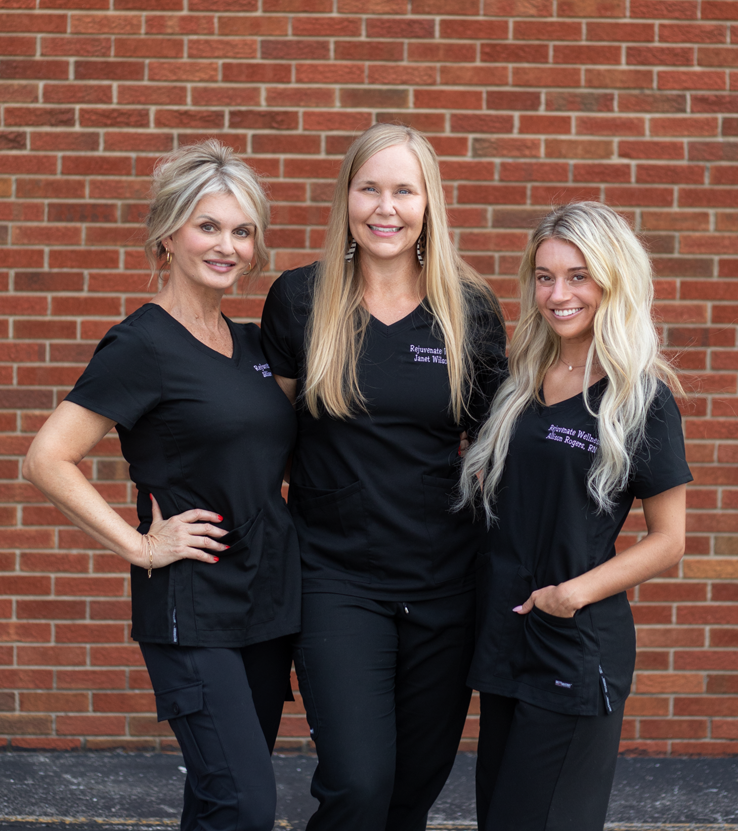Three women in black uniforms standing in front of a red brick wall, smiling at the camera at Rejuvenate Wellness in Florence Kentucky
