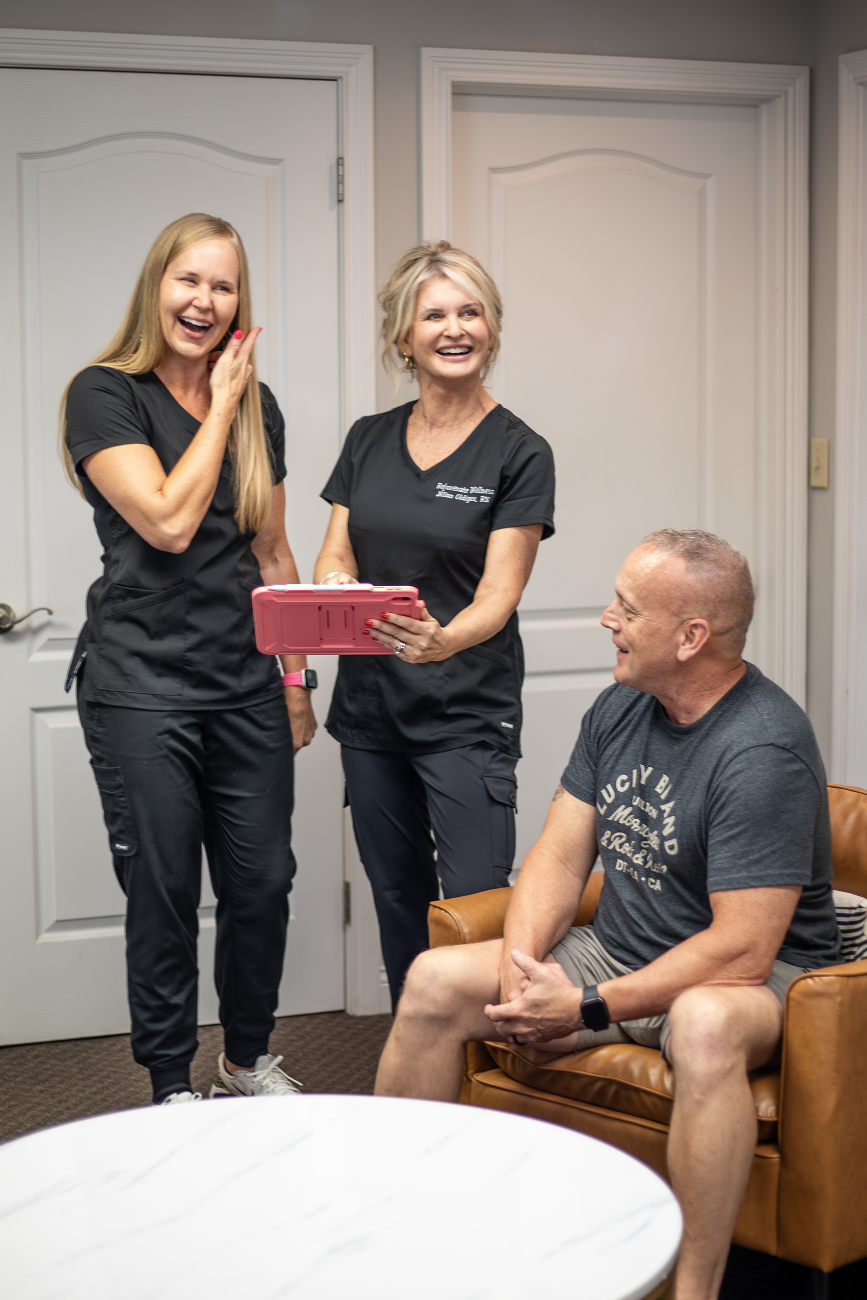 Four people sharing a moment of laughter in a room. Two women in black scrubs standing and smiling, one woman with platinum blonde hair holding a tablet, and a man with a shaved head sitting in a leather chair, also smiling at Rejuvenate Wellness in