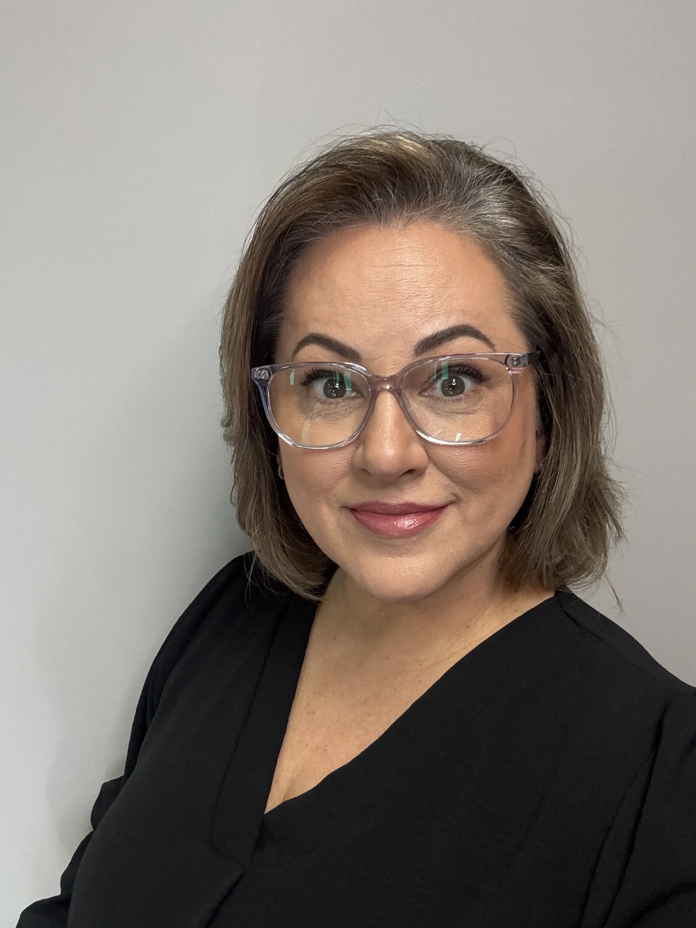 A woman with brown, shoulder-length hair wearing glasses and a black top smiling against a plain gray background at Rejuvenate Wellness in Florence Kentucky