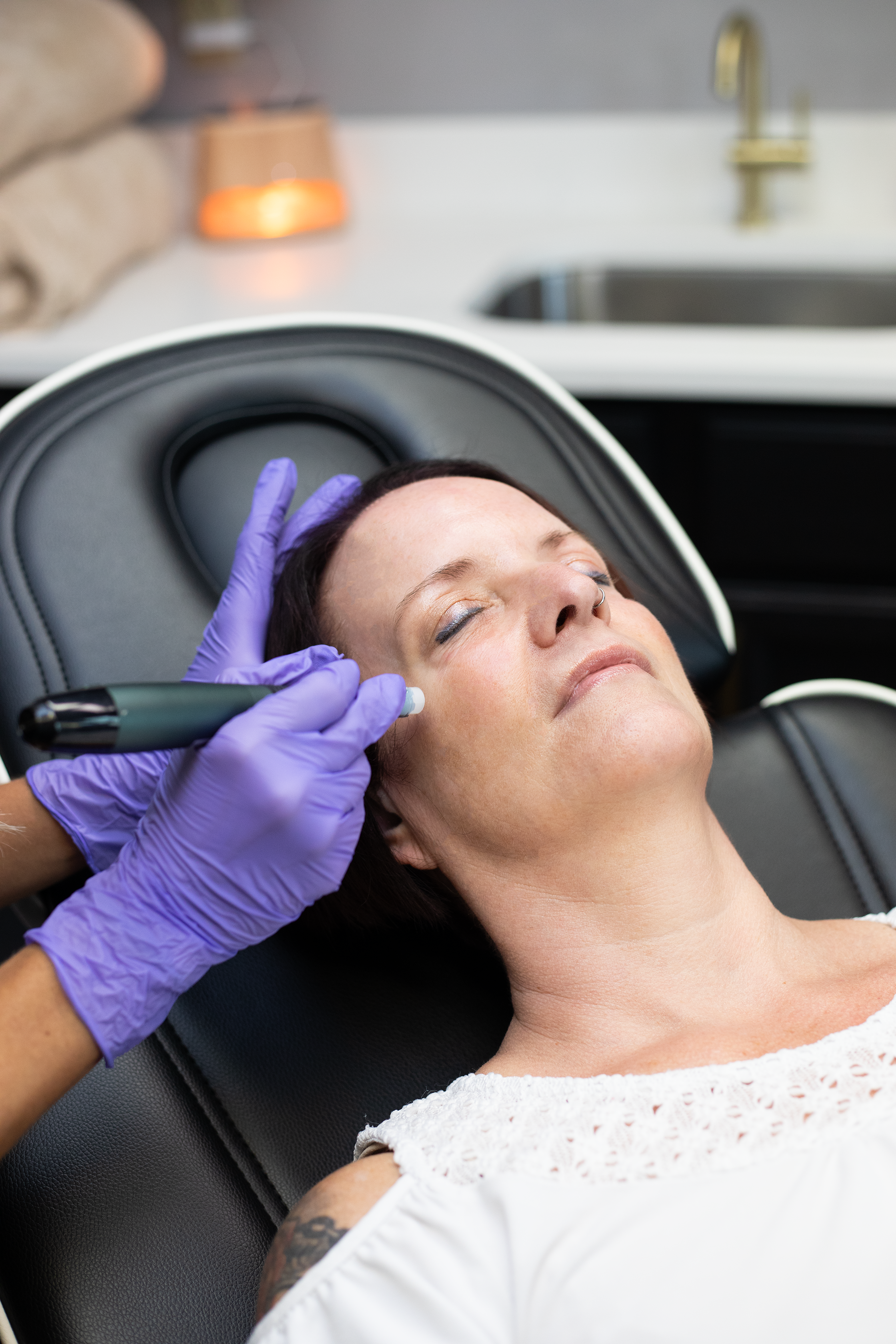 A woman with dark hair and a nose ring is lying on a black reclined chair with closed eyes while a practitioner wearing purple gloves uses a handheld device on her forehead at Rejuvenate Wellness in Florence Kentucky