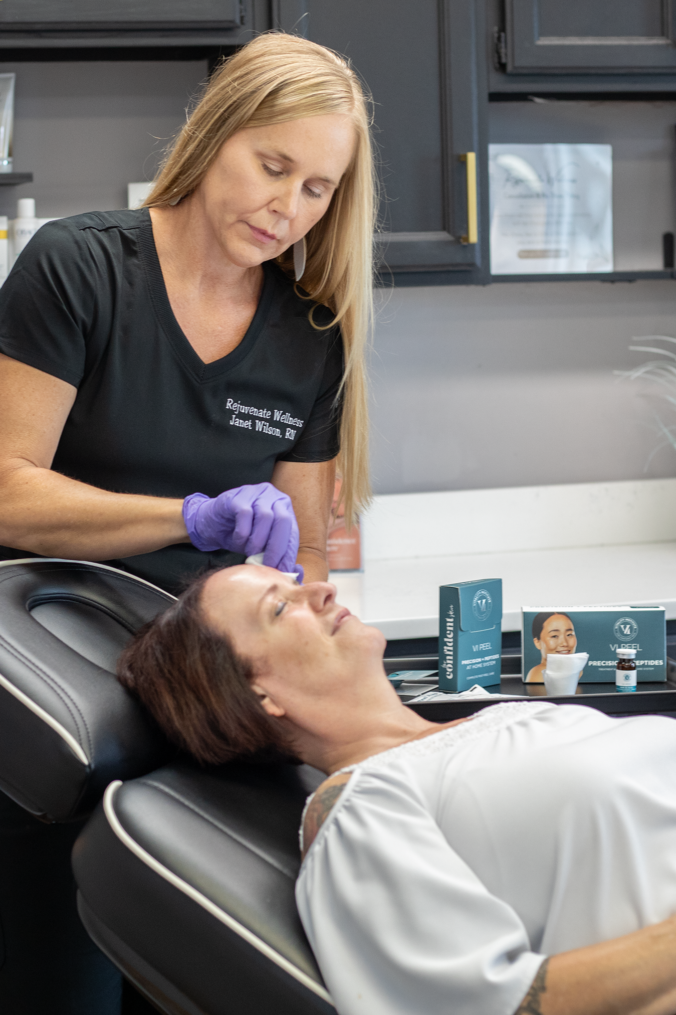 A woman receiving a facial treatment from a skincare professional in a wellness clinic. The professional is wearing a black shirt and purple gloves, and is working on the woman's face. The woman is lying back in a treatment chair with her eyes closed