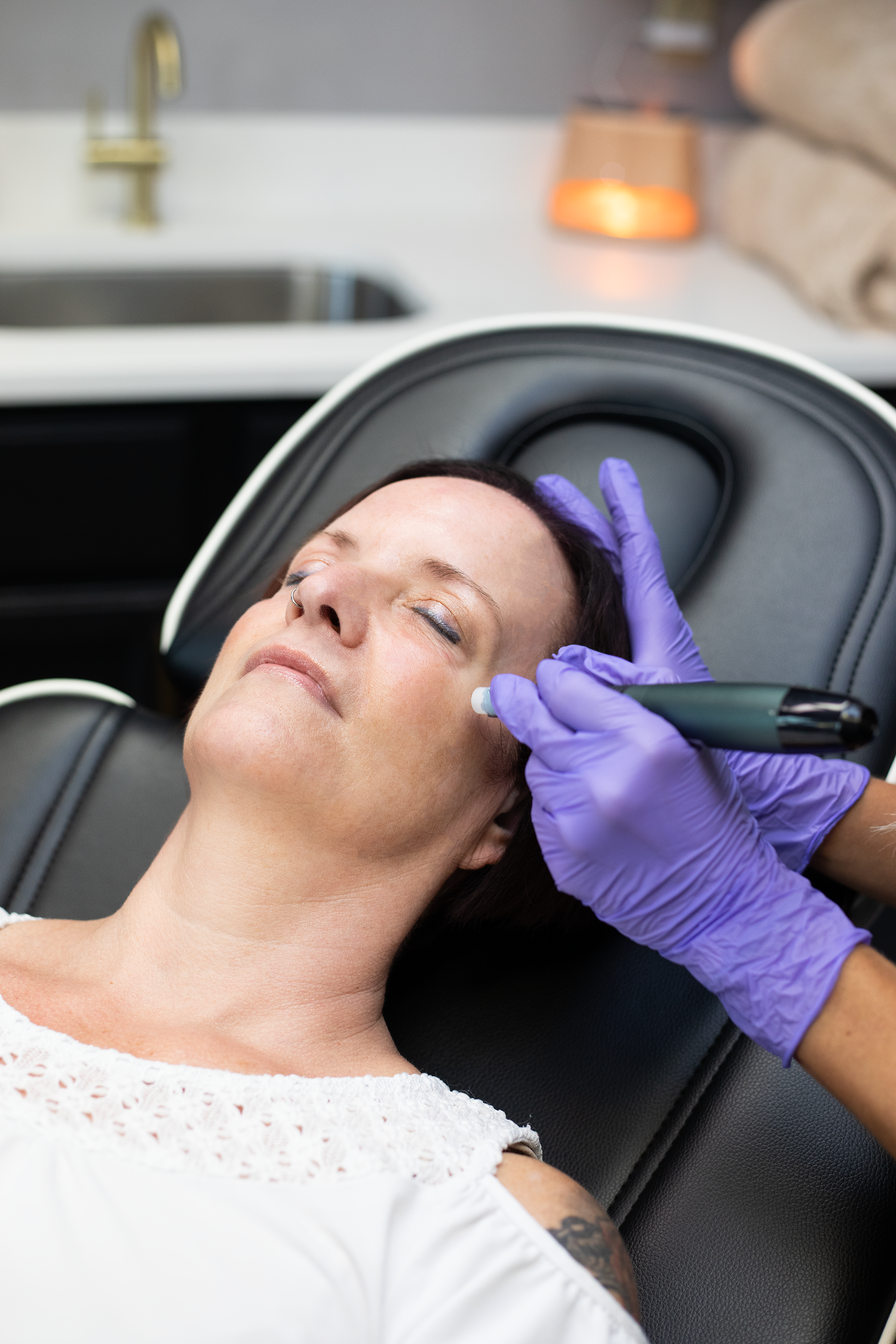 A woman with dark hair and a nose piercing lies back on a reclining chair with her eyes closed as a person wearing purple gloves applies a small device to her face. The background shows a bathroom sink and a lit candle at Rejuvenate Wellness in Flore