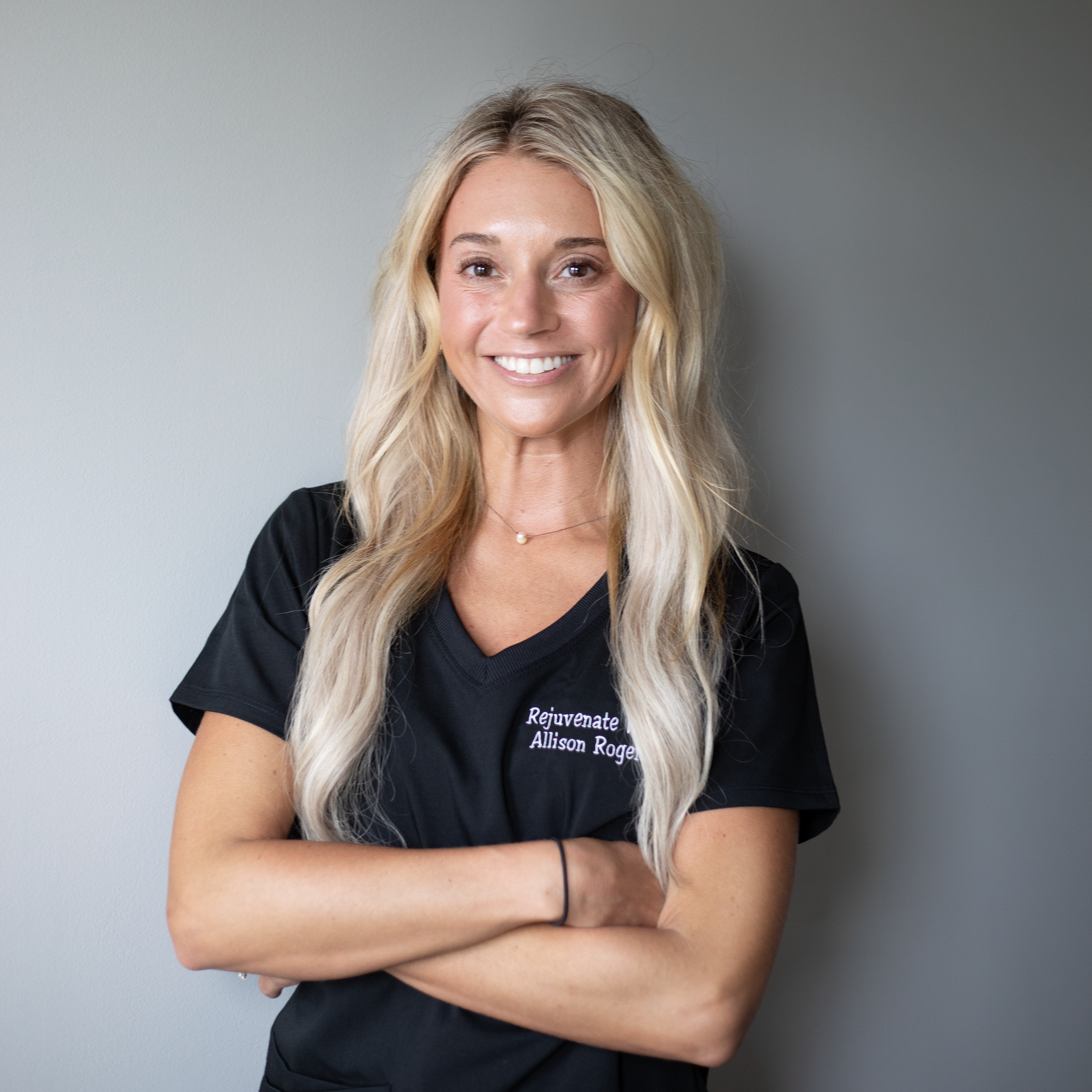 Close-up of a smiling woman with long blonde hair, wearing a black shirt with white text, standing against a plain gray background at Rejuvenate Wellness in Florence Kentucky