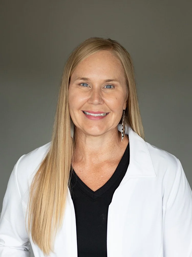 A woman with long blonde hair, blue eyes, and light skin, smiling at the camera. She is wearing a black top, white jacket, and a feather-shaped earring. The background is plain gray at Rejuvenate Wellness in Florence Kentucky