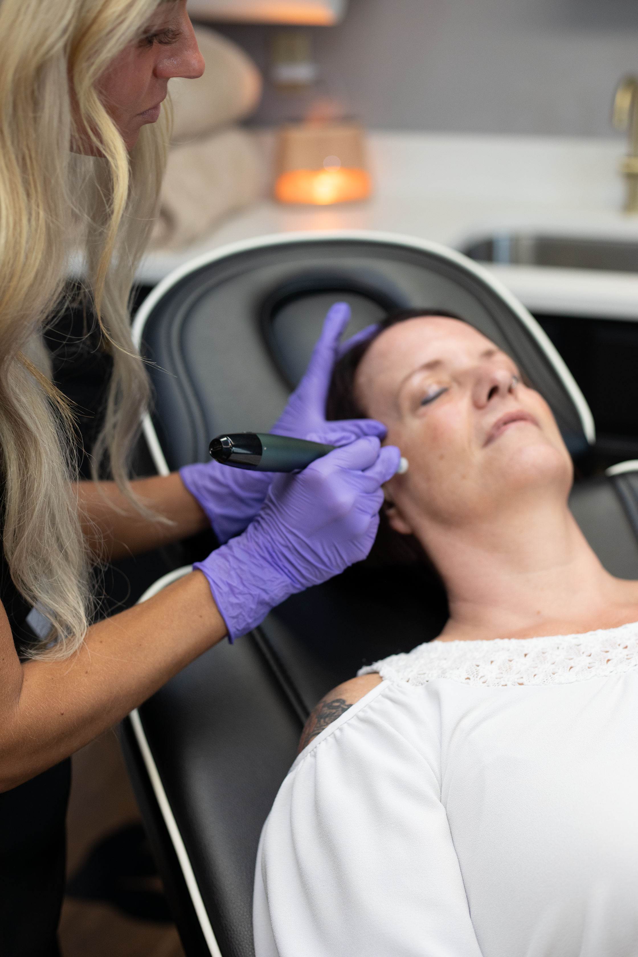 A woman wearing purple gloves is performing a facial treatment with a tool on a patient lying in a reclining chair. The patient has her eyes closed and appears relaxed at Rejuvenate Wellness in Florence Kentucky