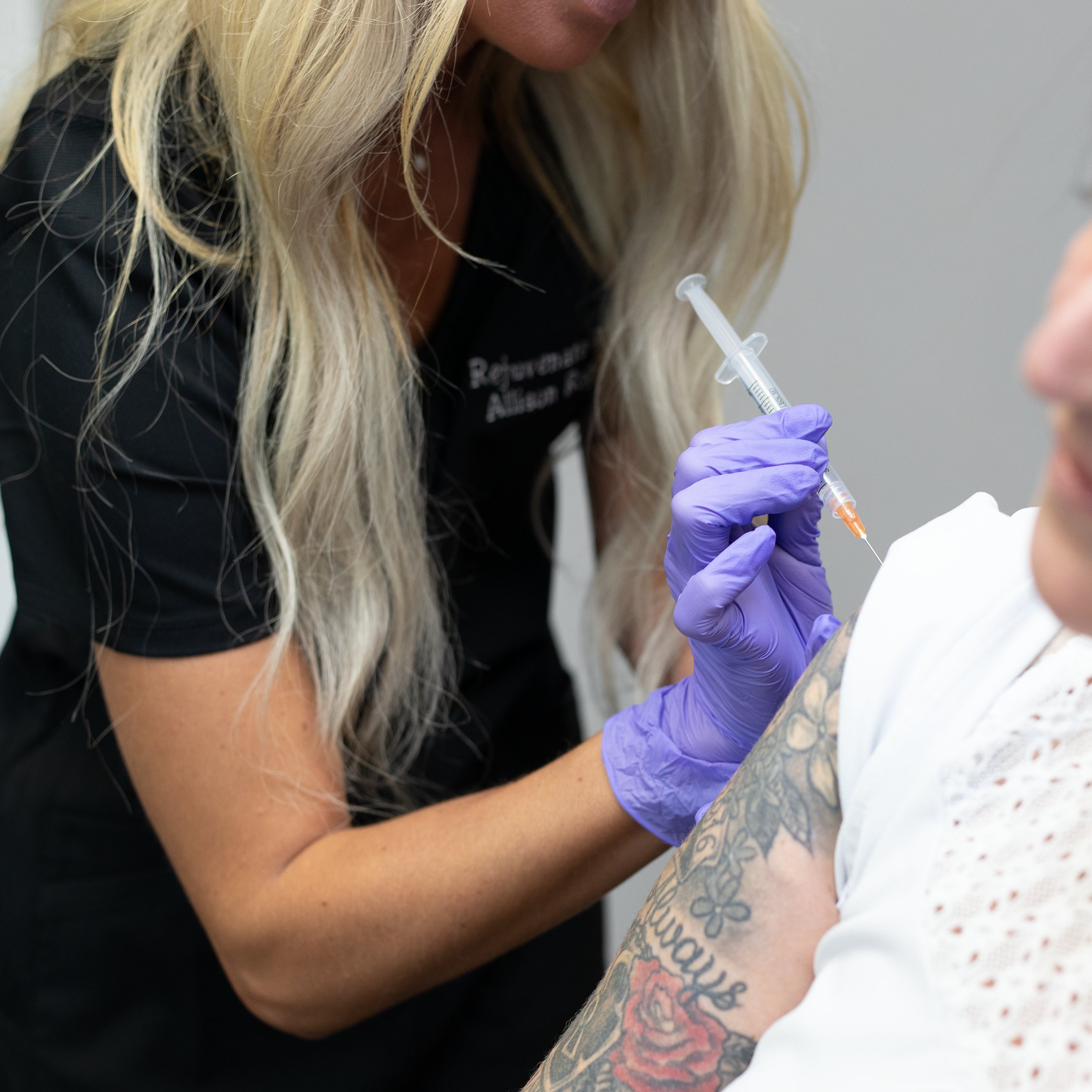 A woman with long blonde hair, wearing a black shirt, is administering a vaccination or injection to a tattooed arm of another person using a syringe with an orange cap, wearing purple gloves at Rejuvenate Wellness in Florence Kentucky