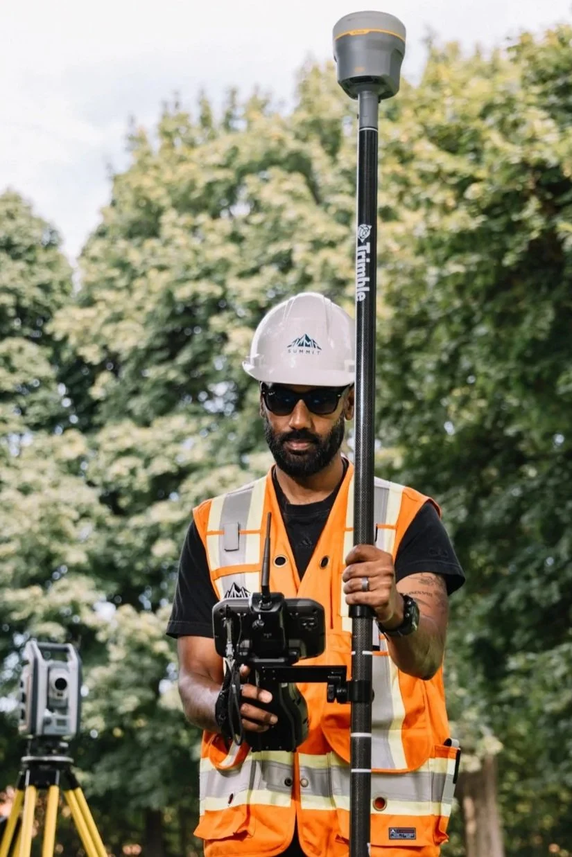 A surveyor wearing a white hard hat, sunglasses, and an orange safety vest holds a GNSS receiver mounted on a pole while working outdoors among trees.
