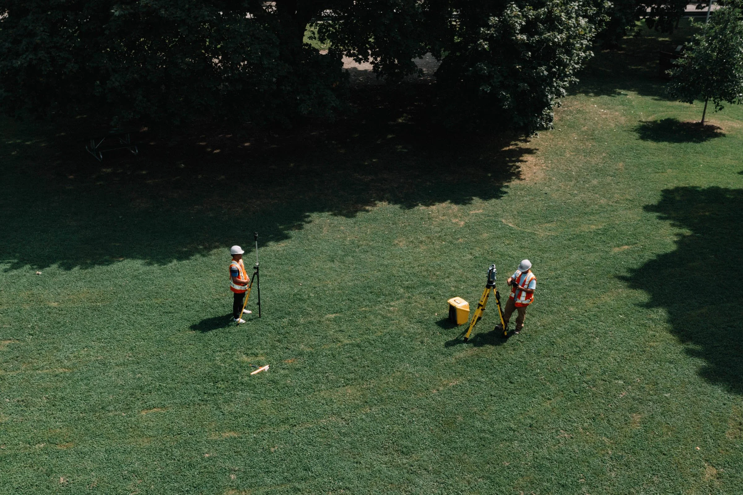 Two workers in safety vests and helmets are conducting field measurements or survey on a grassy park area with trees, using surveying equipment with tripods and a yellow toolbox.